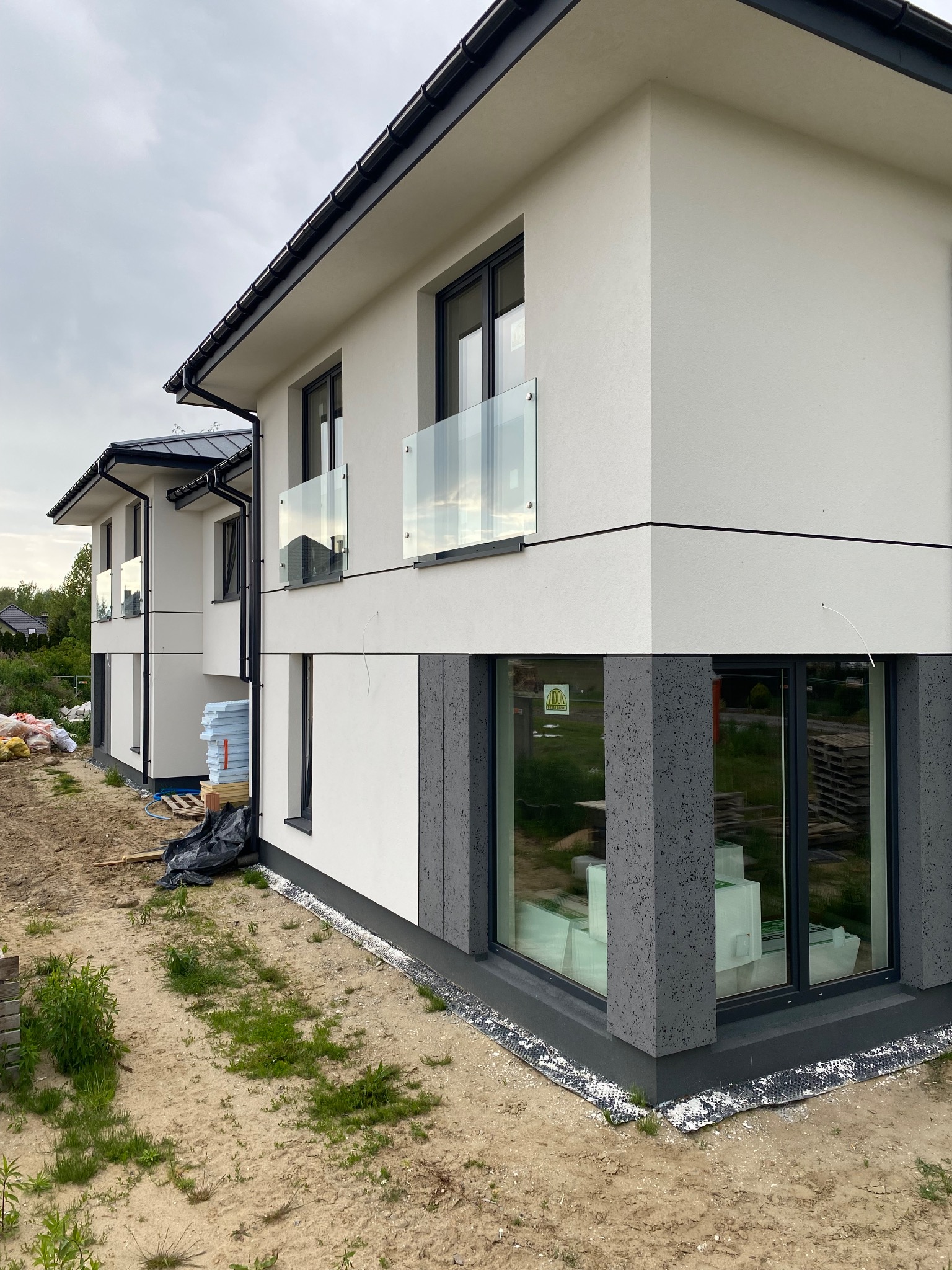 Nowoczesny dom z balkonami z balustradami ze szkła bezpiecznego, duże okna z szarymi ramami i elewacją w odcieniach bieli i szarości. Teren wokół domu w trakcie zagospodarowania.
