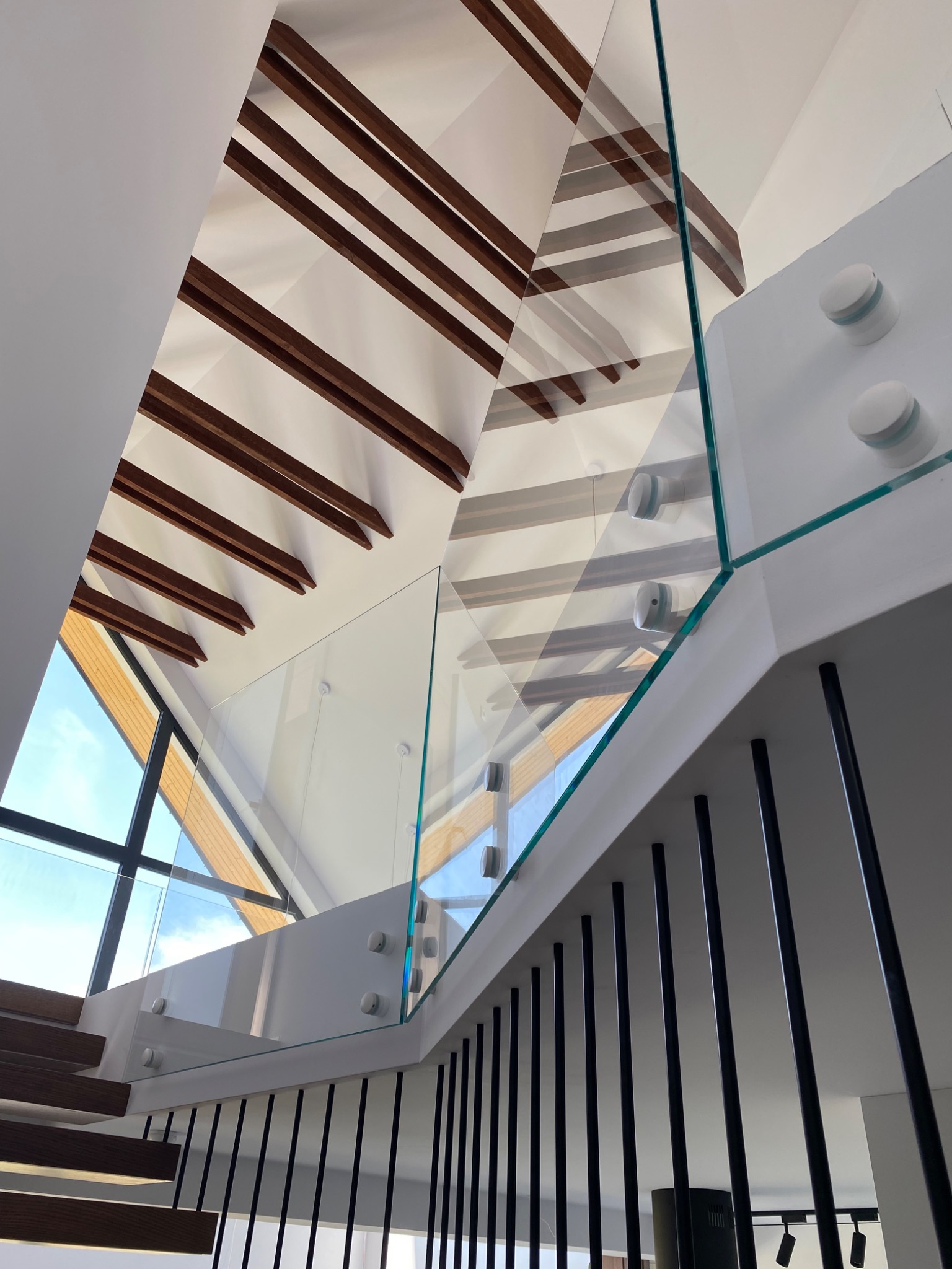 Nowoczesna balustrada schodowa ze szkła hartowanego, mocowana punktowo, z czarnymi, metalowymi elementami. Widok z dołu, na tle drewnianej konstrukcji dachu.