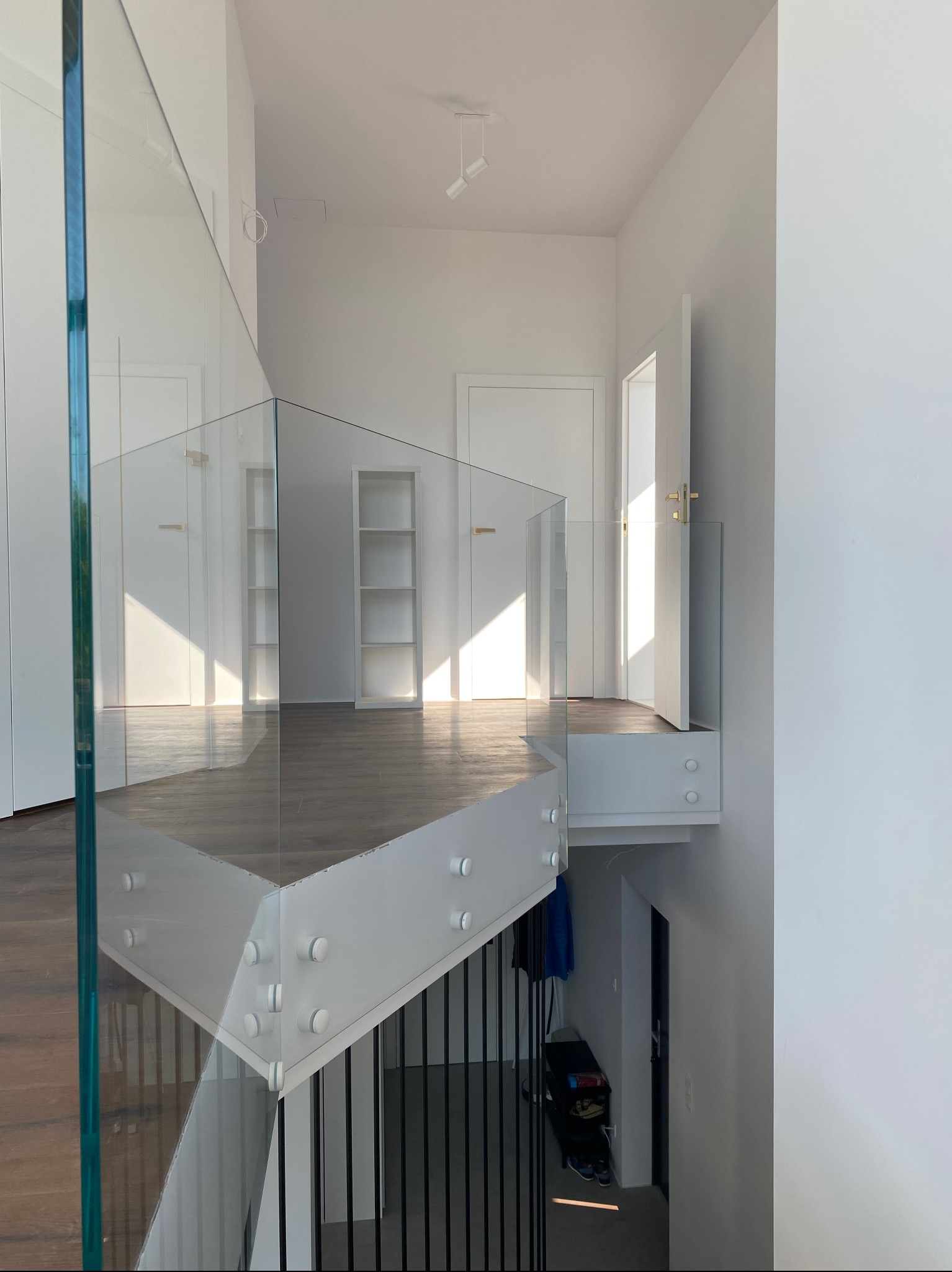 Szklana balustrada schodowa z białymi elementami mocującymi w nowoczesnym wnętrzu. Widoczne schody, drzwi i półka na książki w tle. Minimalistyczny design.