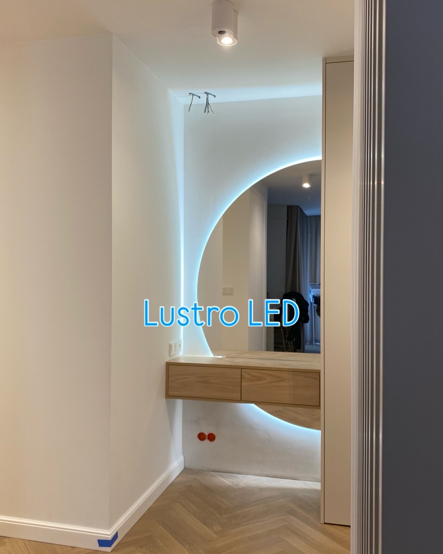 Nowoczesne lustro LED w kształcie półksiężyca, wkomponowane w drewnianą toaletkę, oświetlające jasne wnętrze z parkietem w jodełkę i minimalistyczną zabudową.