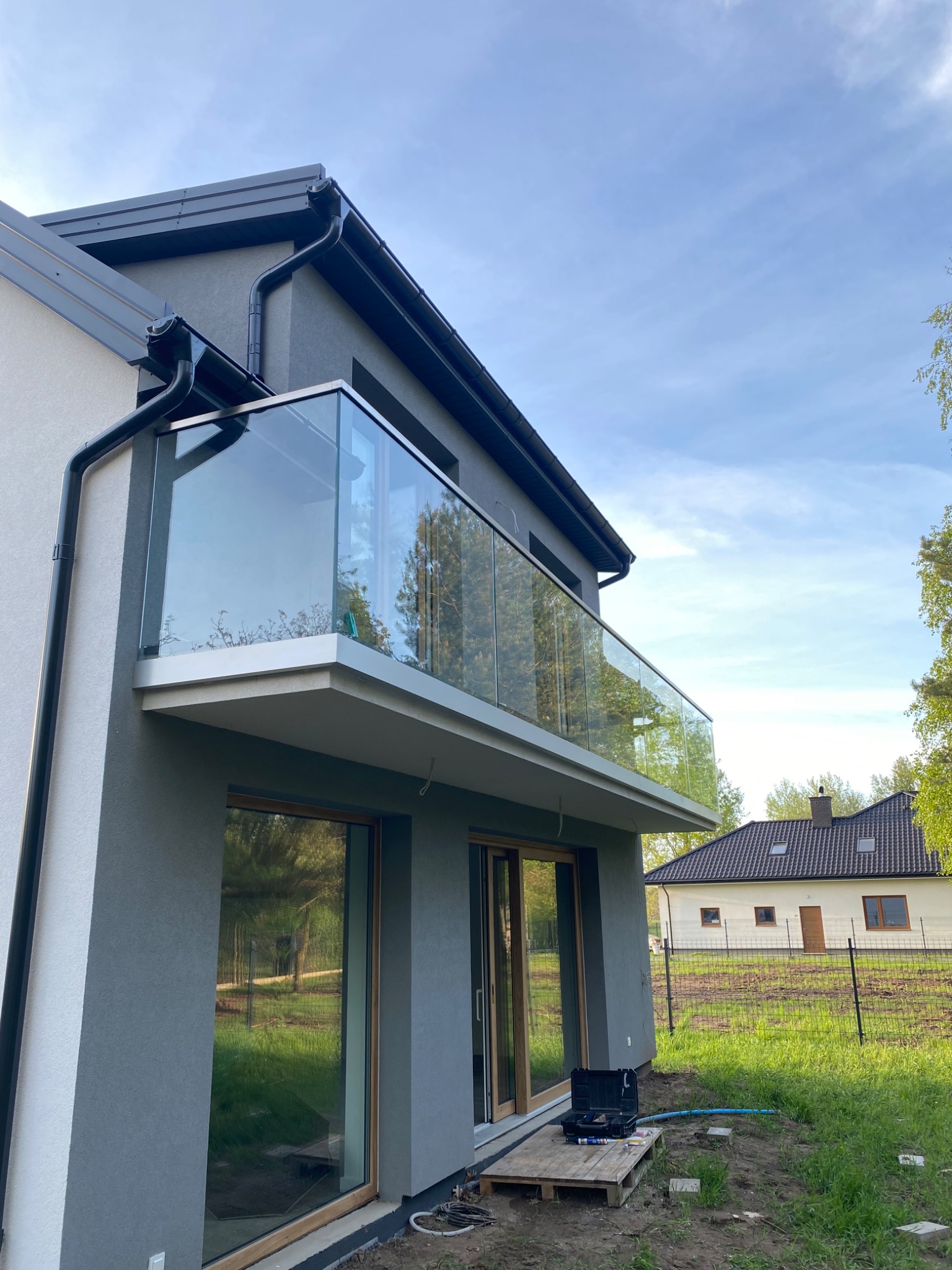 Nowoczesny balkon z szklaną balustradą w domu jednorodzinnym. Widoczne narzędzia i skrzynka monterska sugerują etap końcowy montażu.