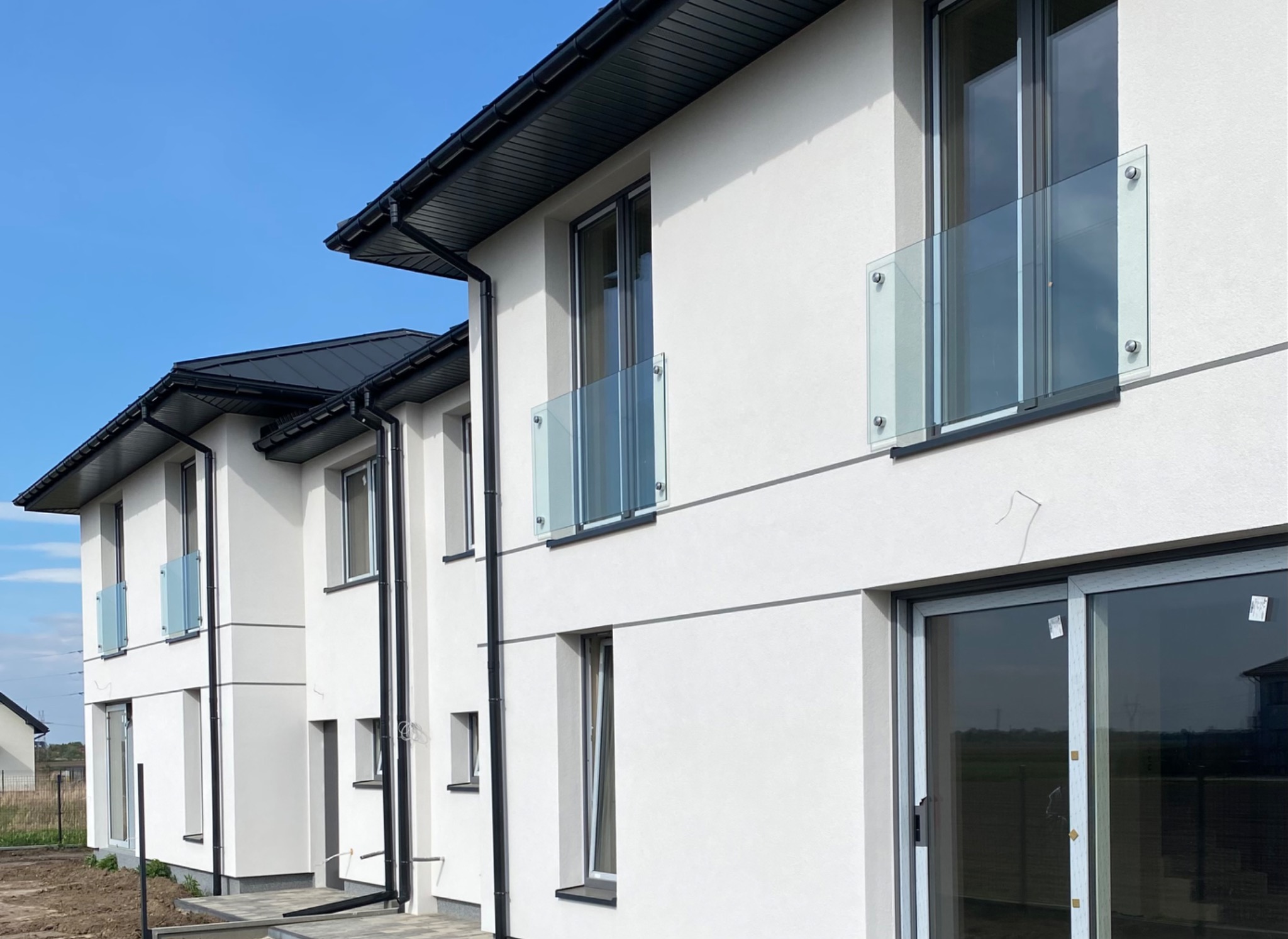 Nowoczesne domy szeregowe z balkonami zabezpieczonymi szkłem, widoczne przesuwne drzwi tarasowe. Minimalistyczna architektura, jasna elewacja, czarne detale.