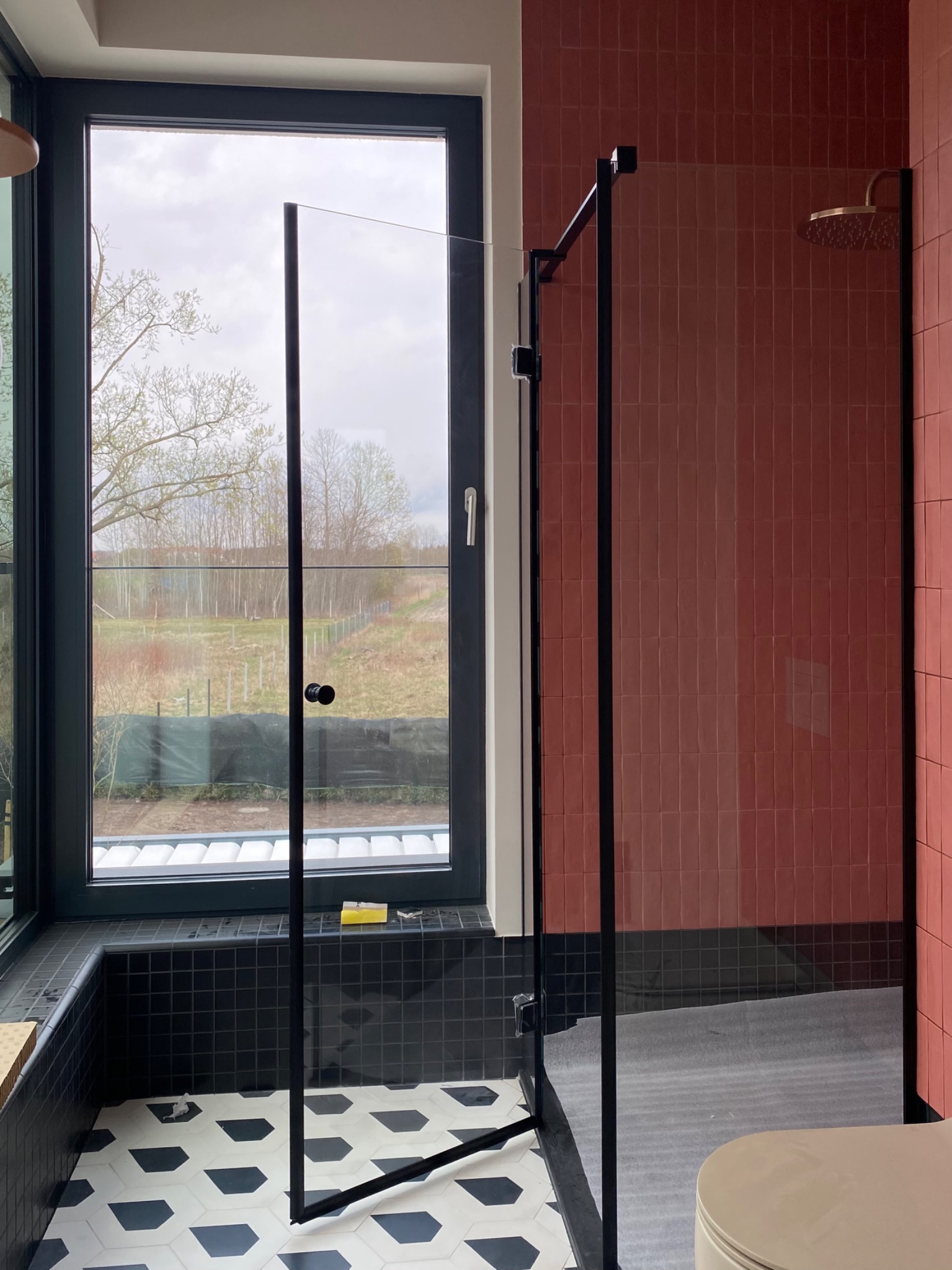Nowoczesna kabina prysznicowa ze szklaną ścianką i czarną ramą na tle okna z widokiem na zewnątrz i czerwonych płytek w łazience. Geometryczne płytki podłogowe.