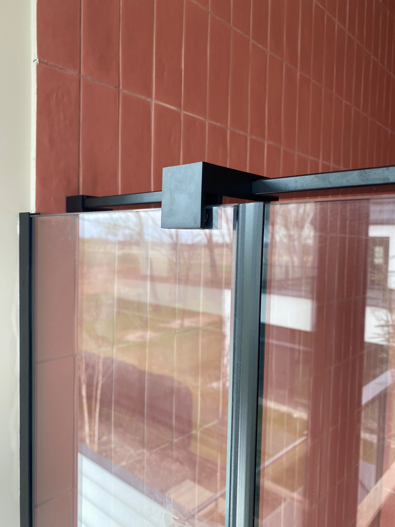 Szklana balustrada z czarnymi elementami mocującymi, widoczna na tle ceglanej ściany. Widok z bliska, ukazujący detale konstrukcji i odbicie otoczenia w szkle.