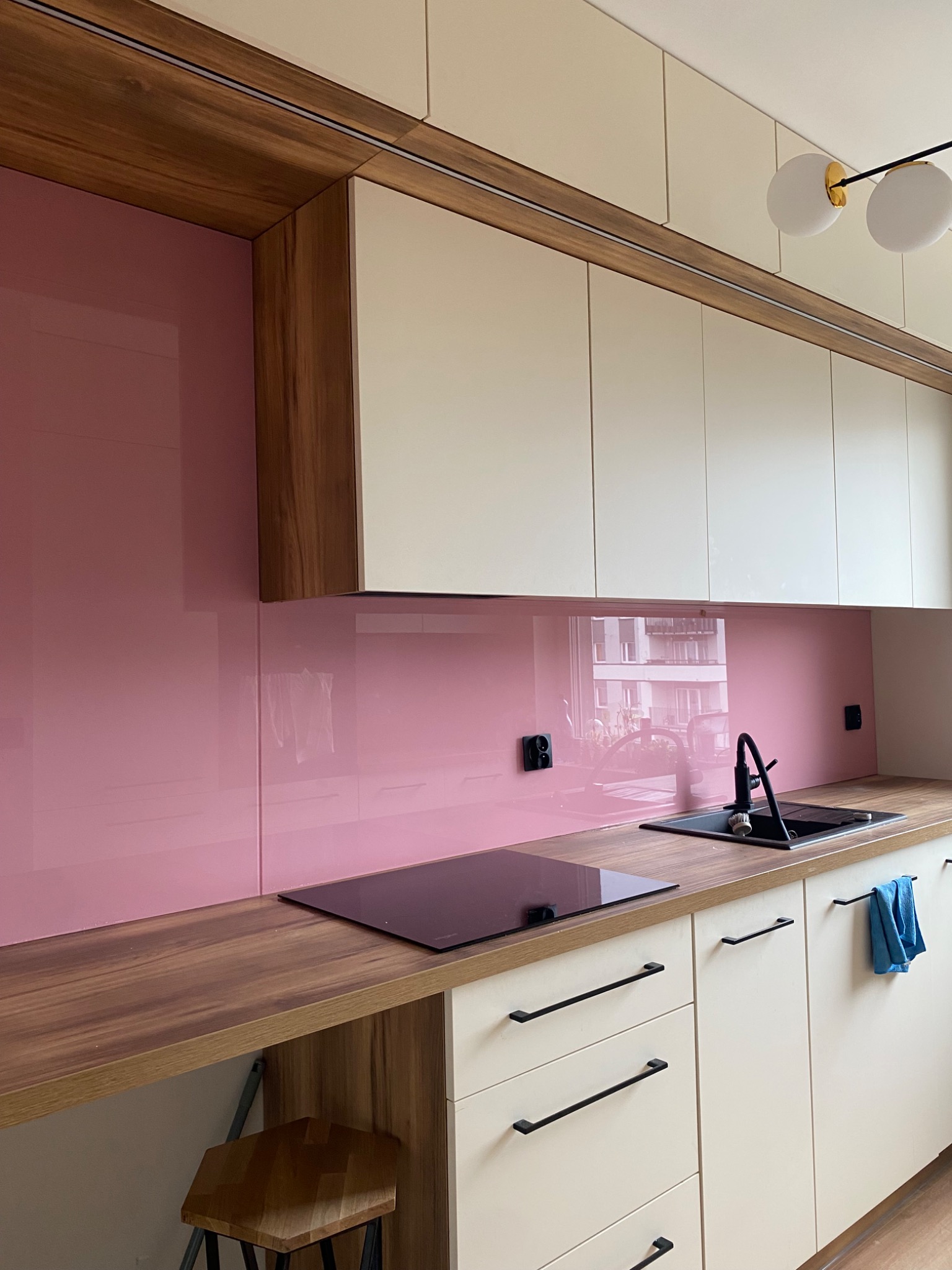 Nowoczesna kuchnia z różowym panelem szklanym nad blatem, drewnianym blatem i białymi szafkami. Widoczny zlew, płyta indukcyjna i minimalistyczne uchwyty.