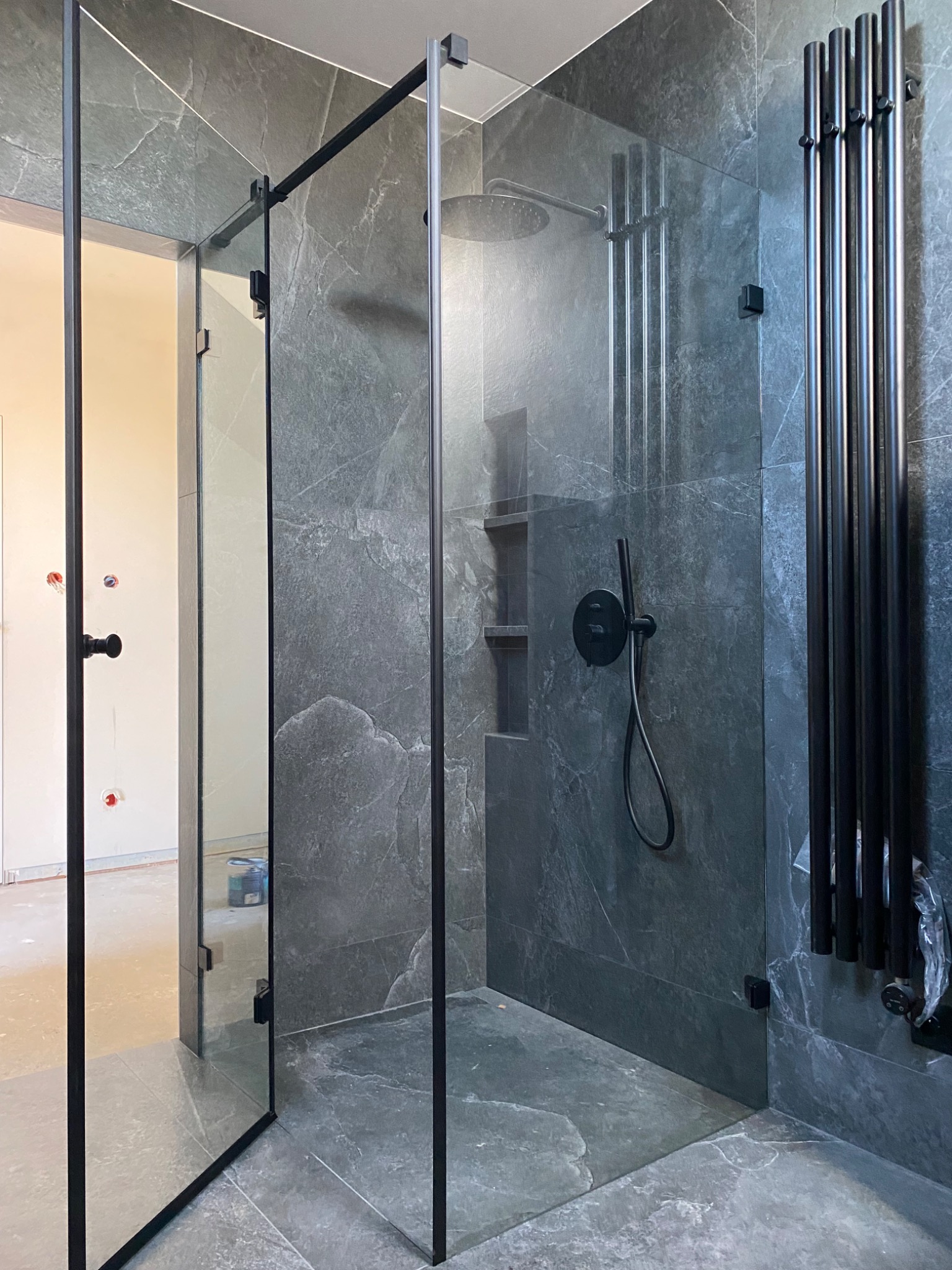Kabina prysznicowa ze szklanymi ścianami i czarnymi profilami na tle szarej, marmurkowej płytki w nowoczesnej łazience. Widoczny grzejnik drabinkowy.