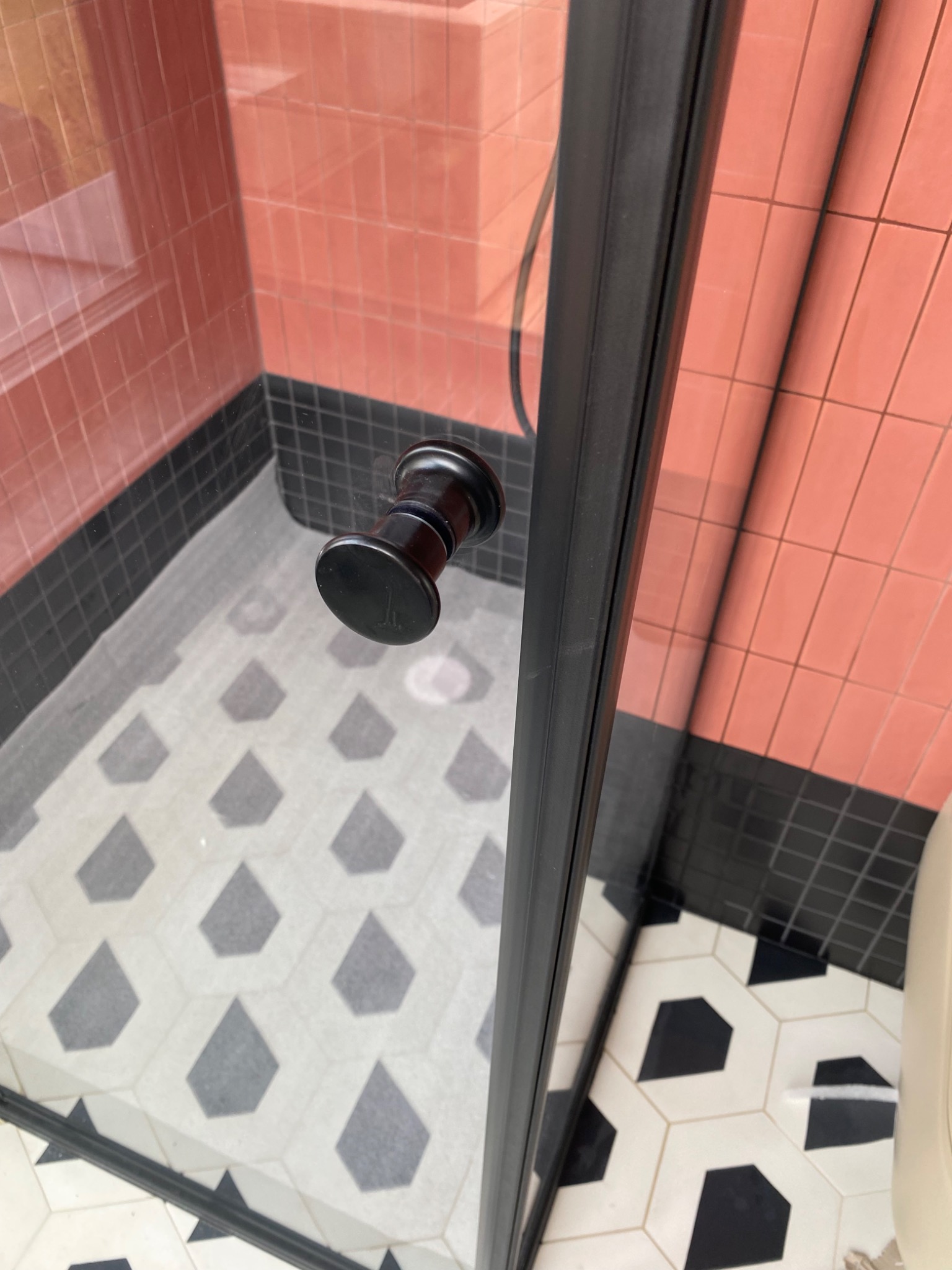 Nowoczesna kabina prysznicowa z czarnymi profilami i okrągłym uchwytem na tle różowych płytek i wzorzystej podłogi.