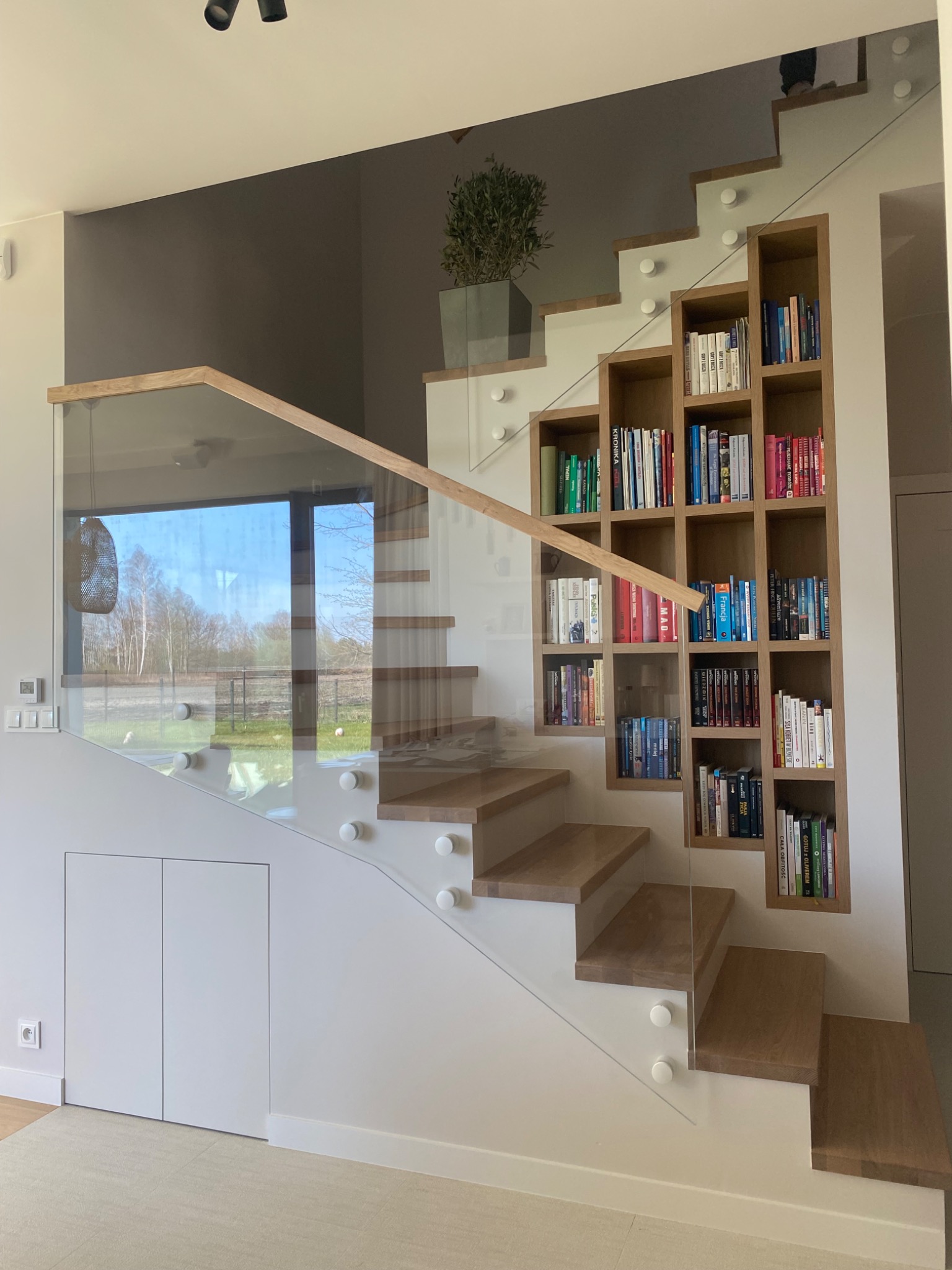 Nowoczesne schody z drewnianymi stopniami i szklaną balustradą, wspornikami punktowymi. Obok schodów regał na książki wbudowany w ścianę.