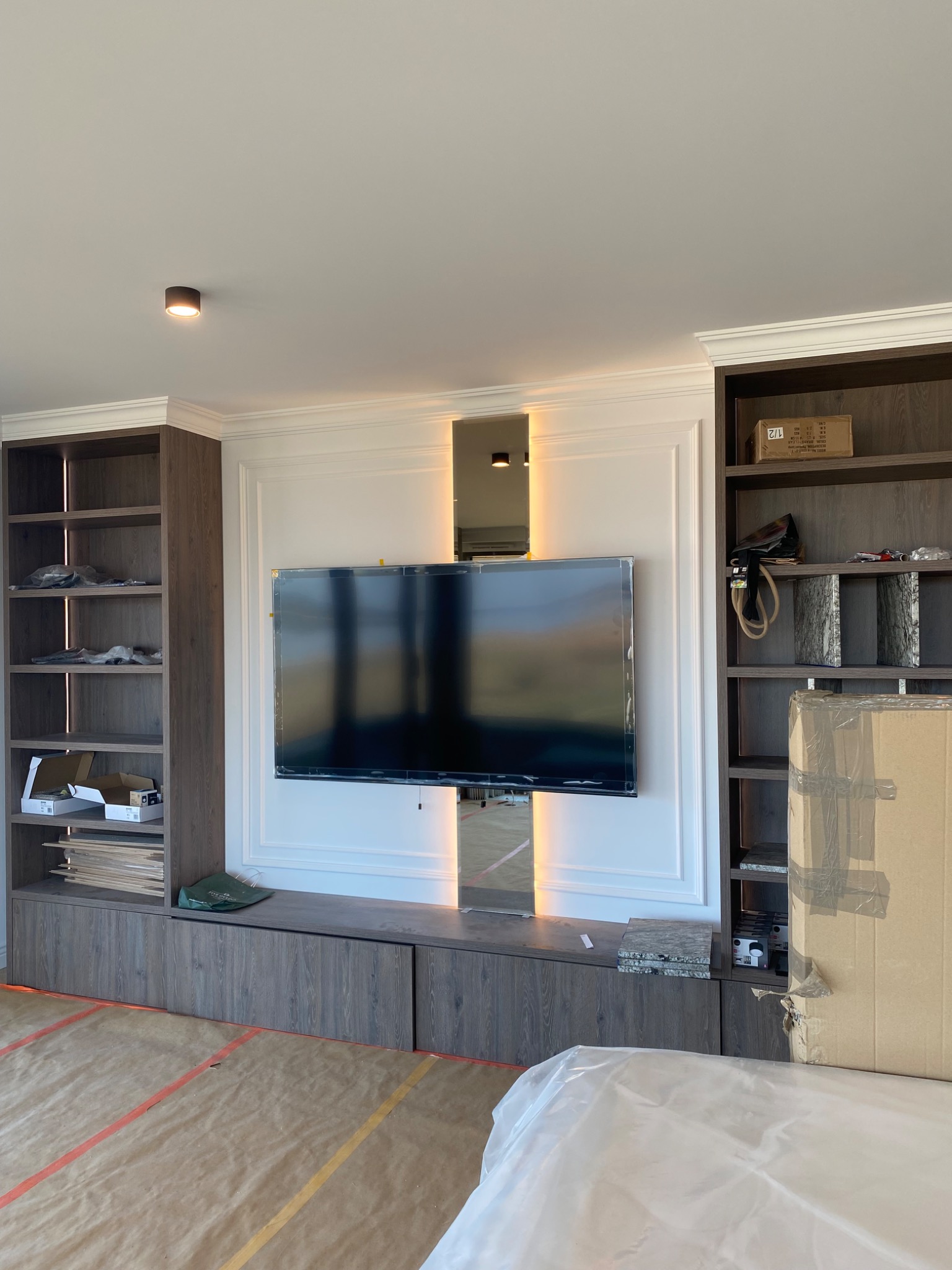 Nowoczesna zabudowa ściany z wnęką na TV i symetrycznie rozmieszczonymi półkami. W tle widoczne lustra i dekoracyjne oświetlenie.