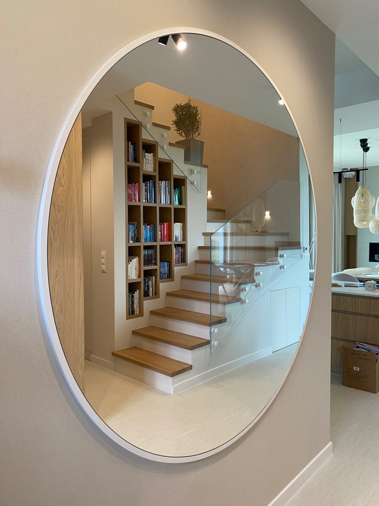 Okrągłe lustro odbija nowoczesne schody z drewnianymi stopniami i szklaną balustradą, obok wbudowany regał na książki. Jasne, minimalistyczne wnętrze.
