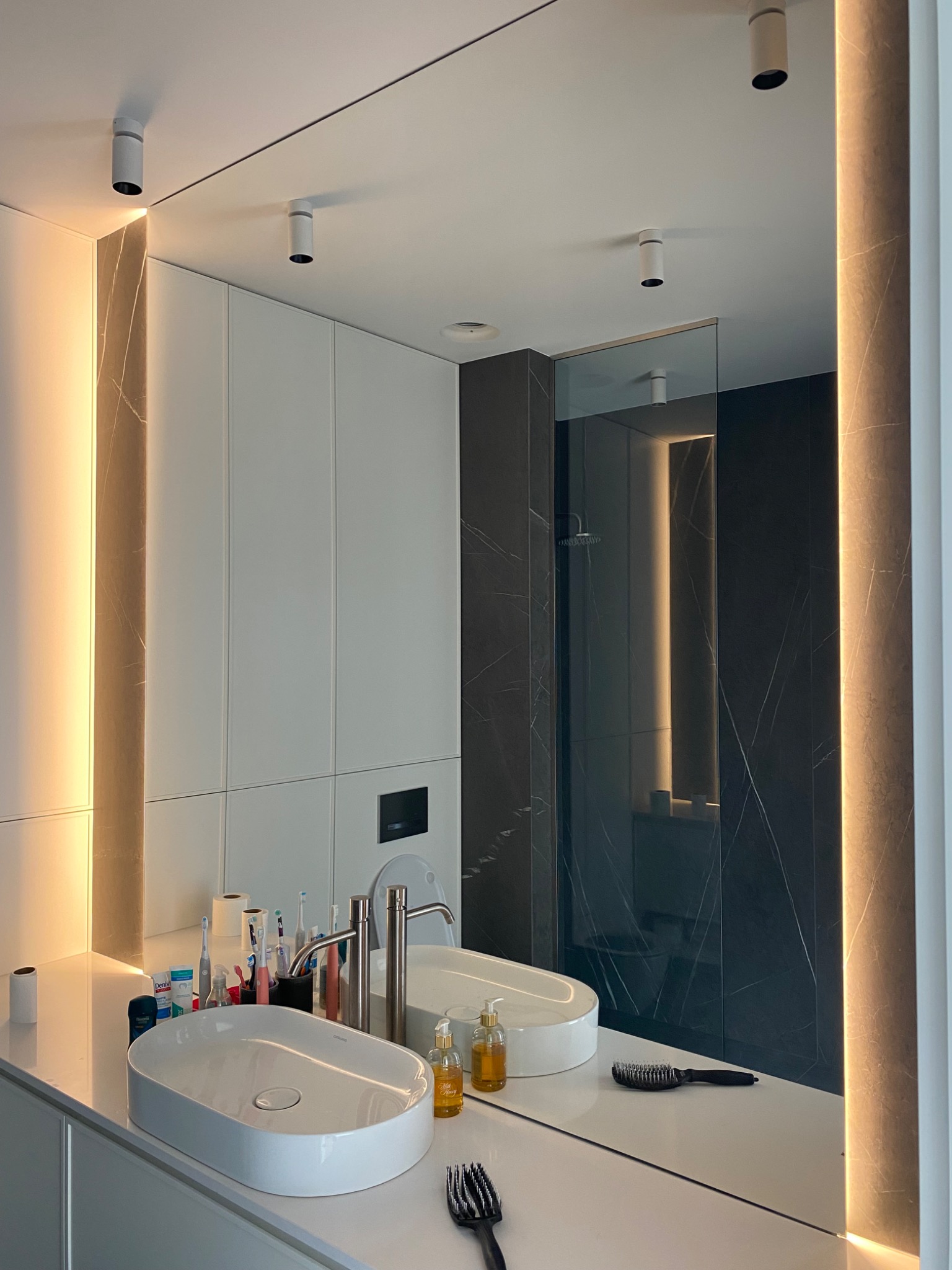 Nowoczesna łazienka z podświetlanym lustrem, białą szafką i umywalką nablatową. Widoczne detale: armatura, oświetlenie punktowe i ciemna ściana prysznicowa.