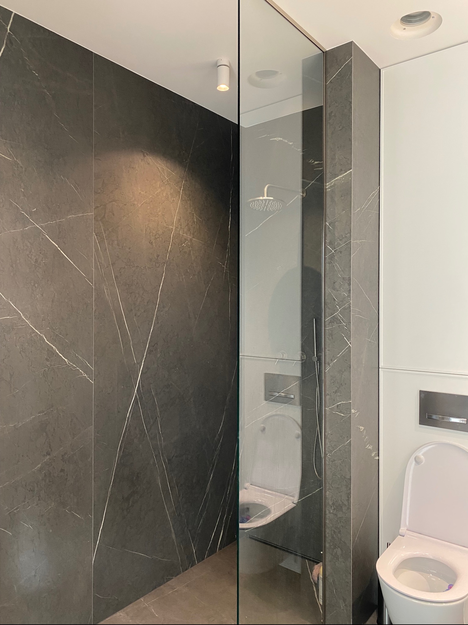 Nowoczesna kabina prysznicowa ze szklaną ścianą, ciemne płytki imitujące marmur z białymi żyłami, minimalistyczna armatura, toaleta w tle.