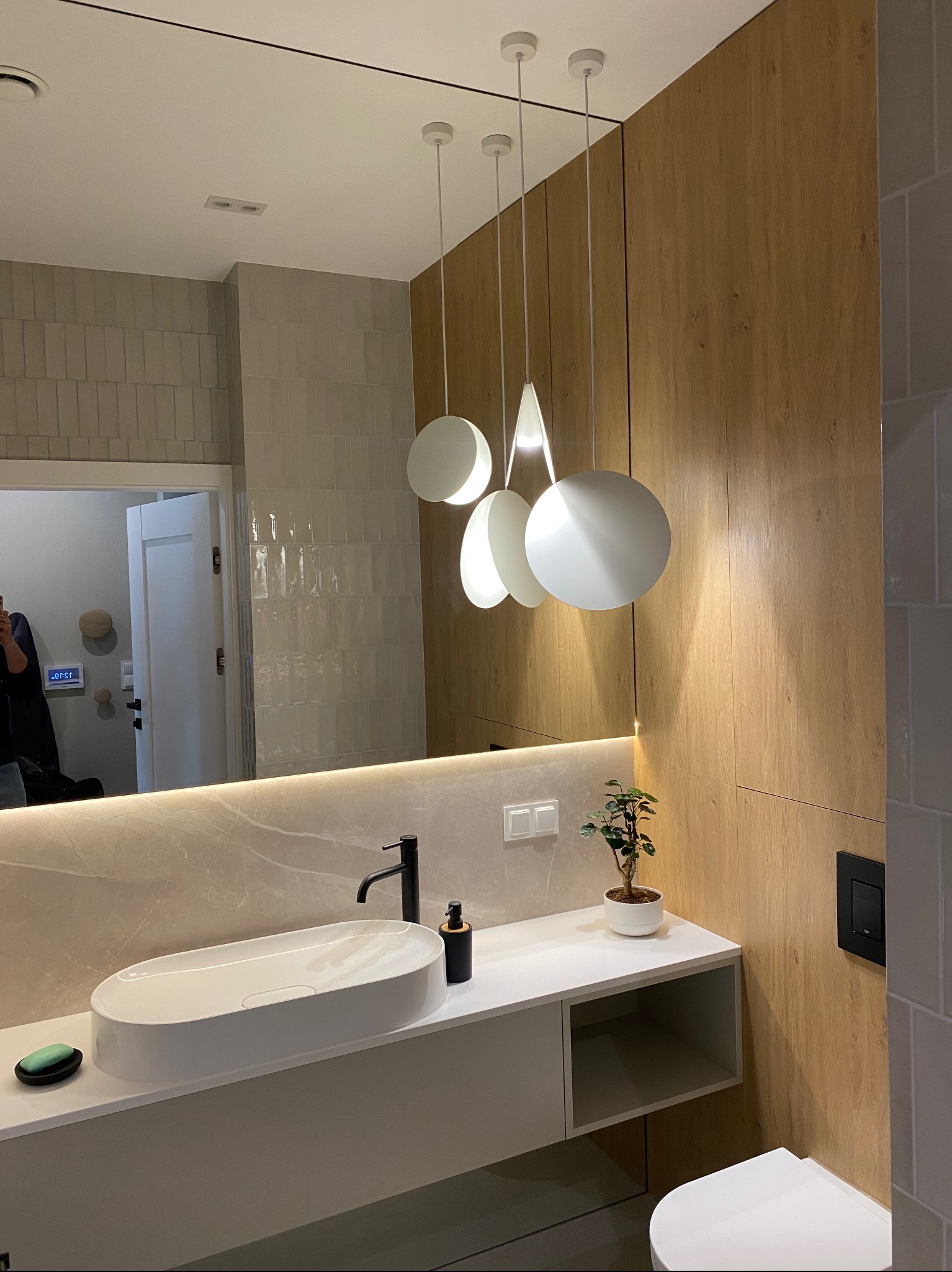 Nowoczesna łazienka z owalną umywalką nablatową, minimalistyczną armaturą i lustrem na całą ścianę, oświetlonym wiszącymi lampami. Widoczne drewniane panele ścienne.