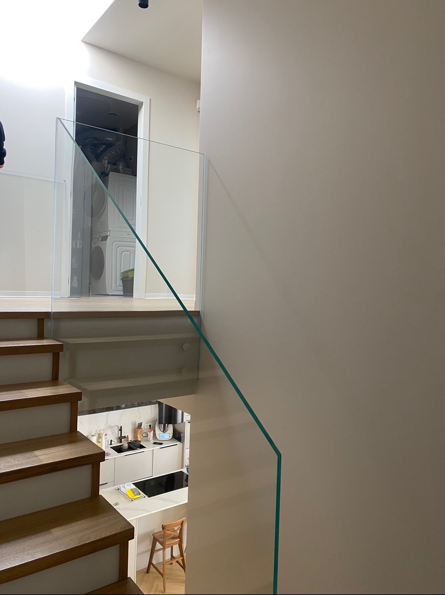 Szklana balustrada na schodach z drewnianymi stopniami, widok z góry na kuchnię i pralkę w tle. Minimalistyczny design, jasne wnętrze.