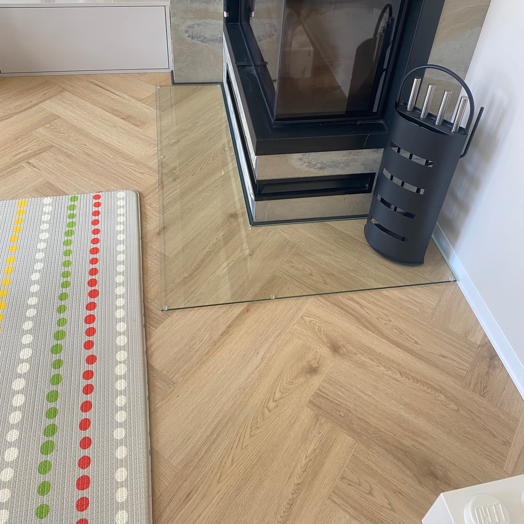 Szklana podłoga pod kominkiem, zabezpieczająca drewniany parkiet ułożony w jodełkę. Obok czarny stojak na narzędzia kominkowe i mata dla dzieci w kropki.