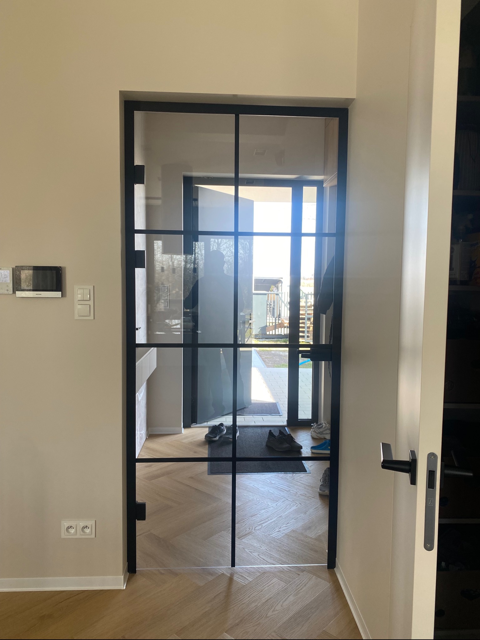 Szklane drzwi w czarnej ramie, widok z wnętrza domu na zewnątrz. Podłoga z parkietem w jodełkę, w tle widoczne buty i wycieraczka.