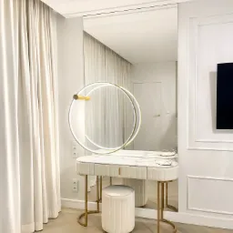 Toaletka w bieli i złocie z okrągłym lustrem podświetlanym LED, na tle jasnych zasłon i parkietu ułożonego w jodełkę.