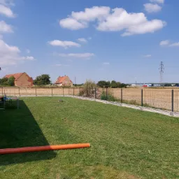 Nowoczesne ogrodzenie panelowe z podmurówką prefabrykowaną na trawniku, w tle budynki gospodarcze i słupy wysokiego napięcia.