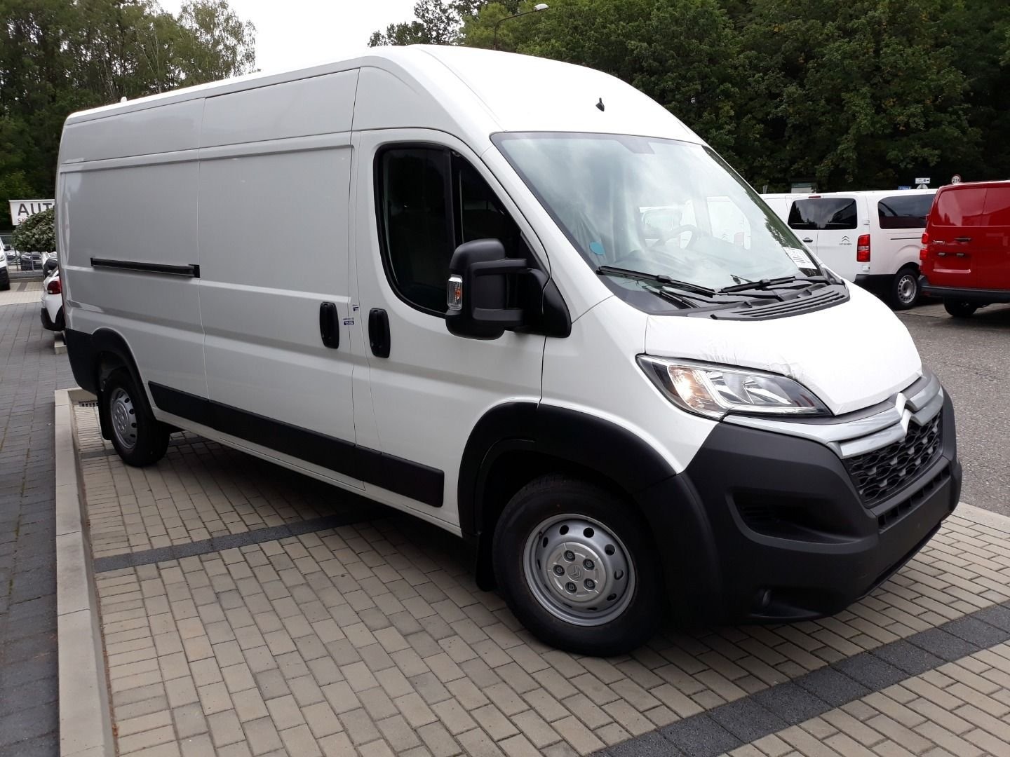 Biały samochód dostawczy Citroen Jumper zaparkowany na brukowanym placu, widok z przodu i boku, z innymi pojazdami w tle.