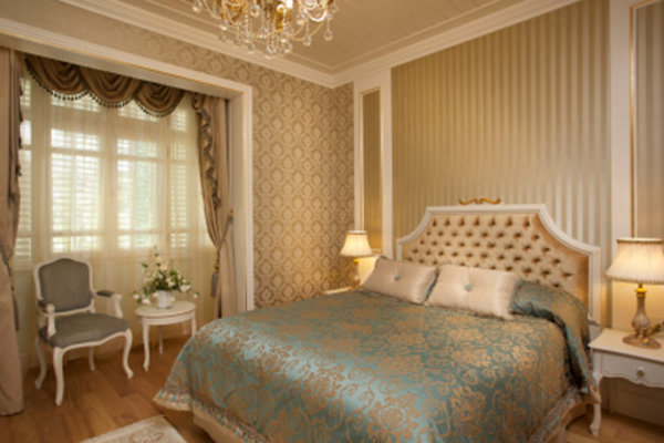 Elegancka sypialnia w stylu klasycznym z wzorzystą tapetą, kryształowym żyrandolem, łóżkiem z pikowanym zagłówkiem i turkusową narzutą, oraz fotelem w stylu Ludwika XVI przy oknie.