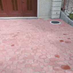 Ułożona czerwona kostka brukowa przed budynkiem z brązowymi drzwiami i szarą elewacją, widoczny właz kanalizacyjny i pionowy element dekoracyjny na fasadzie.