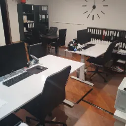 Biuro z białymi biurkami, komputerami, czarnymi krzesłami obrotowymi, regałami z segregatorami i zegarem ściennym bez cyfr.