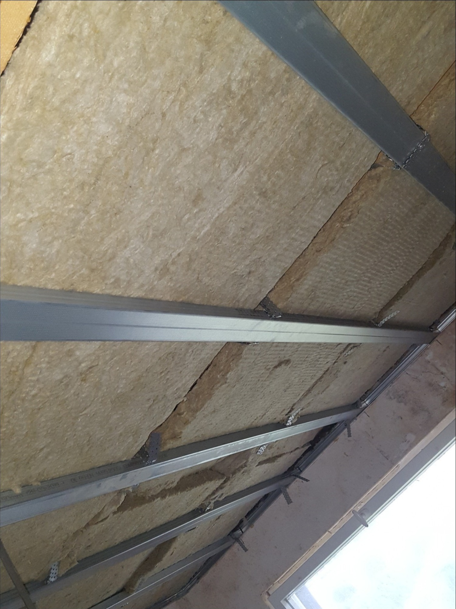 Izolacja poddasza wełną mineralną zamocowaną pomiędzy metalowymi profilami, widoczne okno dachowe.