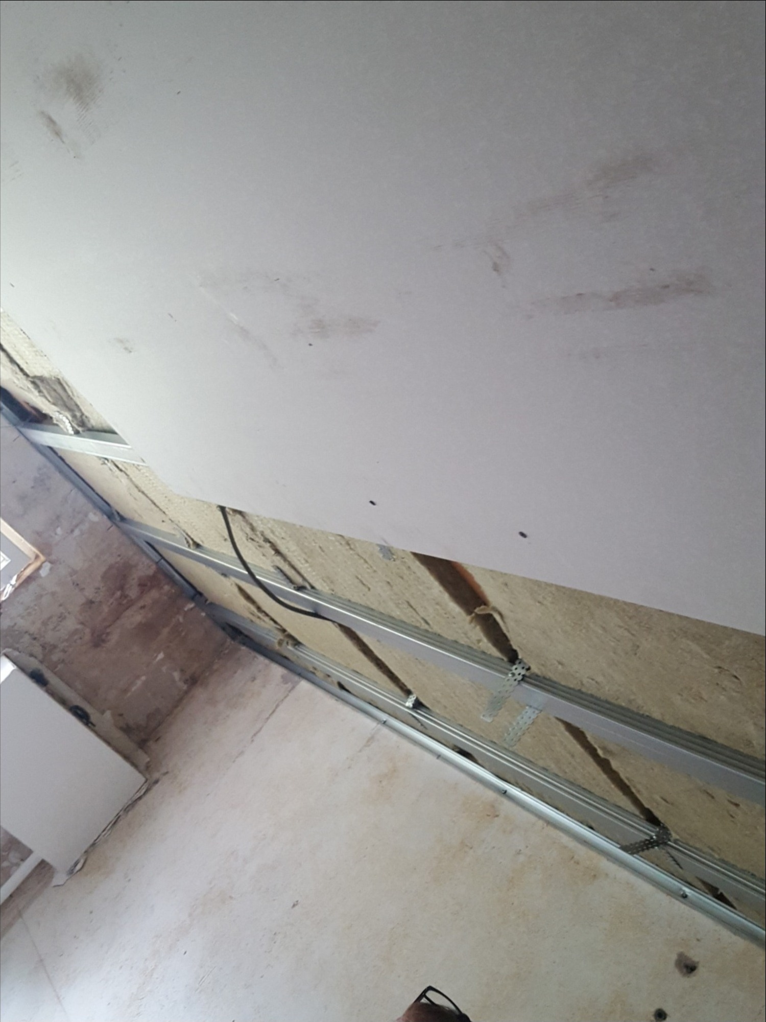 Montaż sufitu podwieszanego z płyt gipsowo-kartonowych na metalowej konstrukcji, widoczne ocieplenie z wełny mineralnej, surowy stan wykończenia ścian.