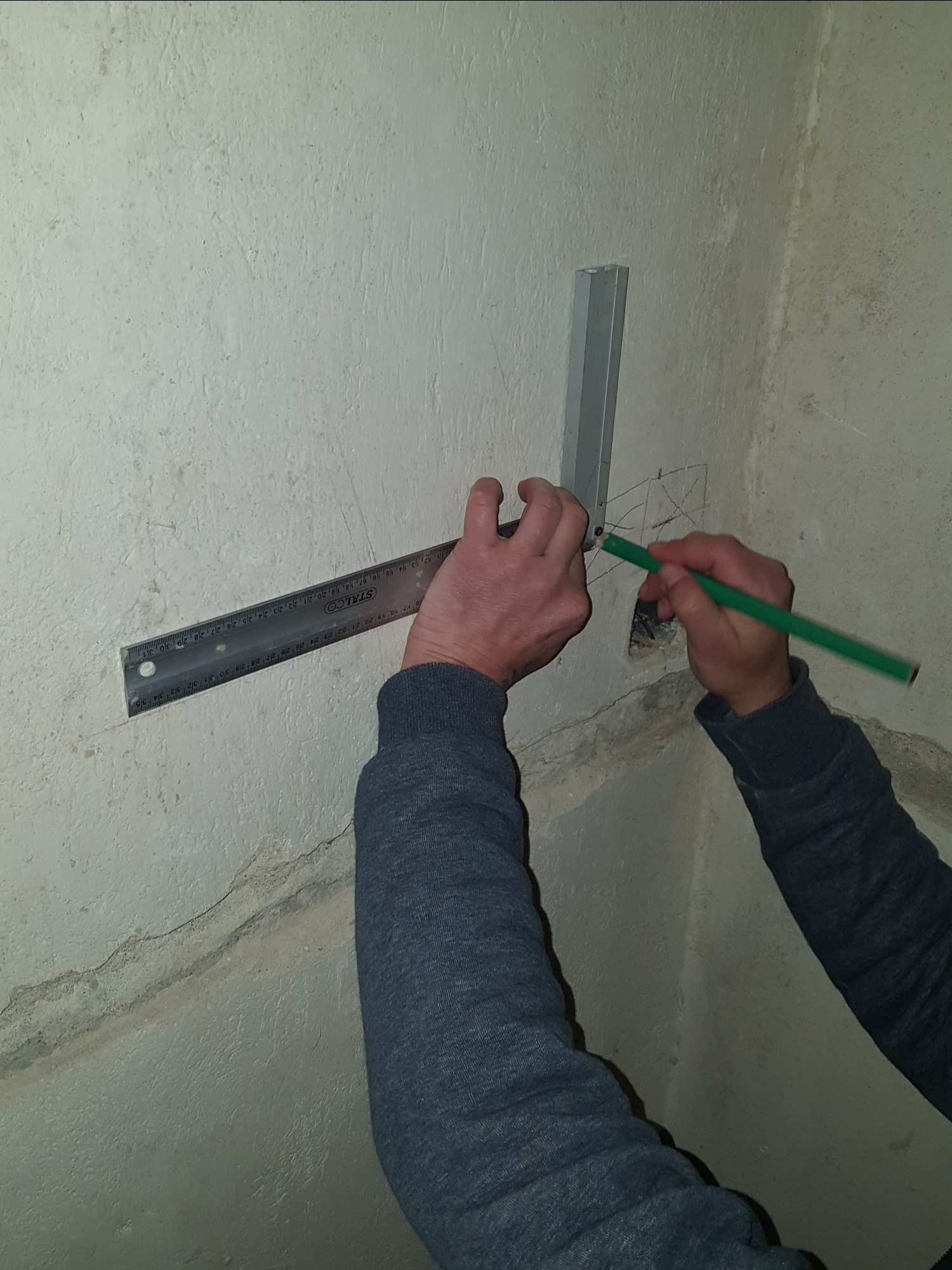 Osoba zaznacza ołówkiem miejsce na ścianie przy pomocy kątownika i metalowej listwy, przygotowując się do dalszych prac wykończeniowych.