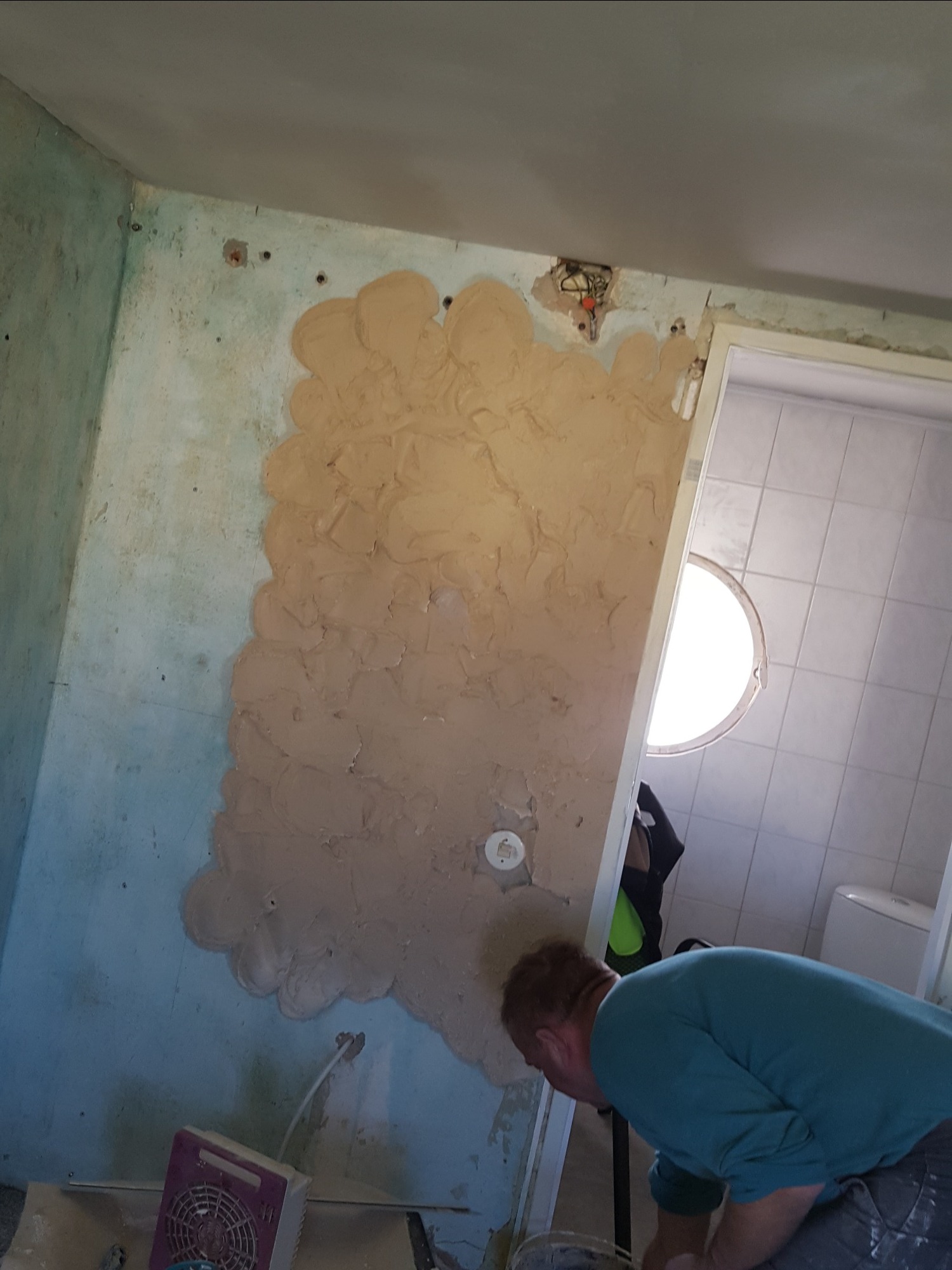 Fragment ściany w trakcie prac przygotowawczych do wykończenia, widoczne nałożone warstwy szpachli, przygotowanie do malowania, w tle widoczna łazienka z okrągłym oknem.