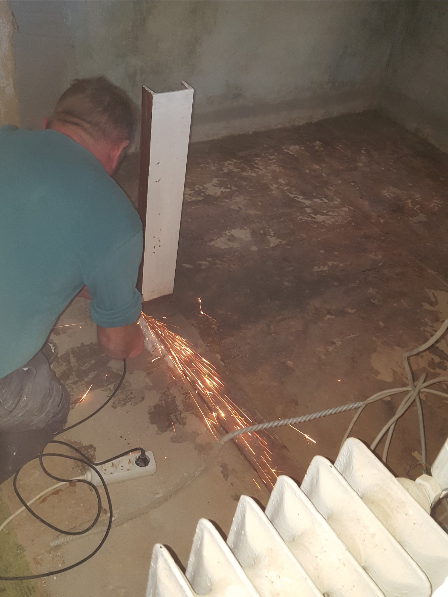 Pracownik w trakcie cięcia metalowego profilu w pomieszczeniu z widocznym grzejnikiem i przedłużaczem, iskry rozchodzą się na boki.