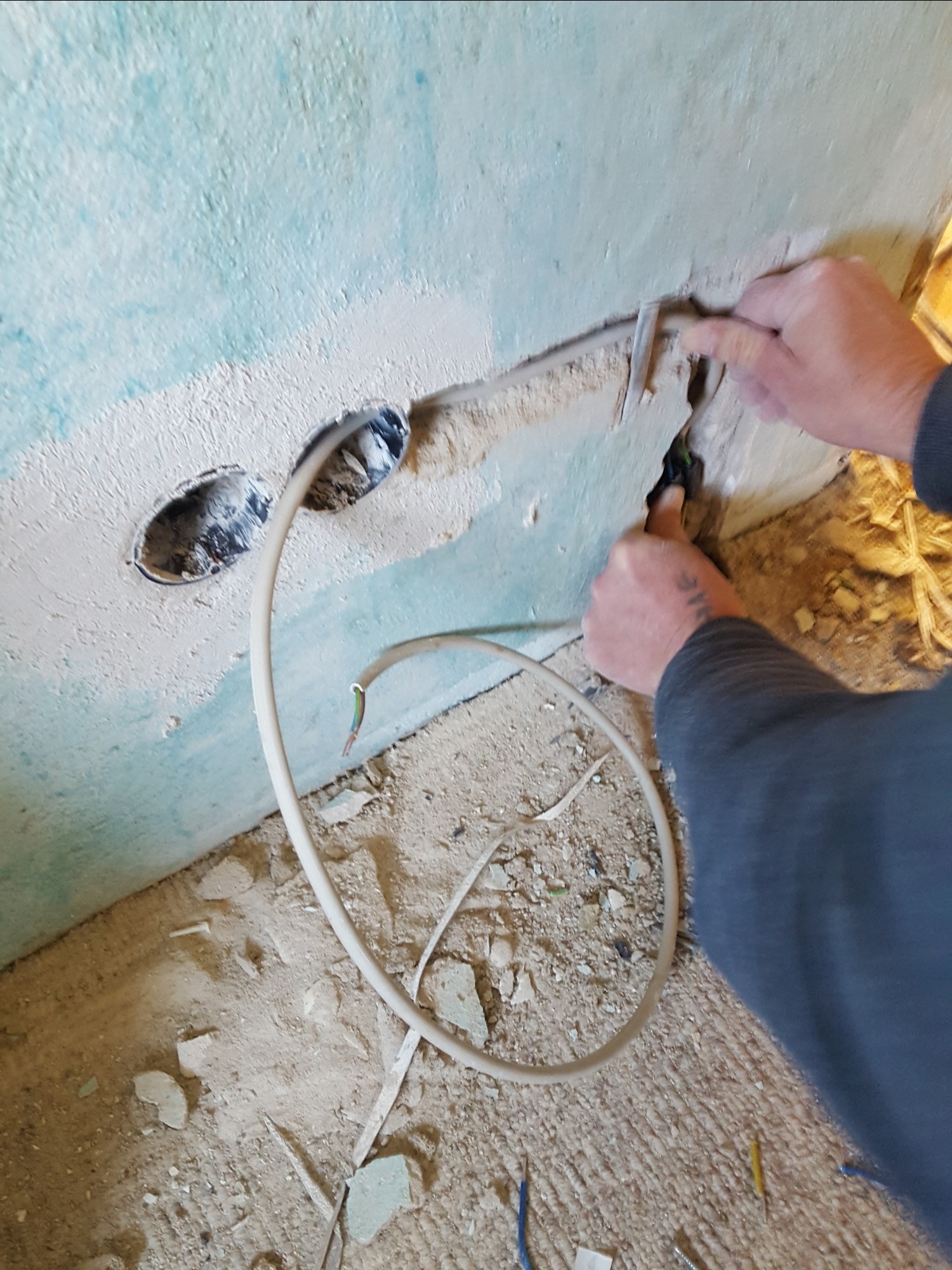 Elektryk z tatuażem na przedramieniu łączy przewody w ścianie z wywierconymi otworami, widoczne kable i gruz na podłodze.