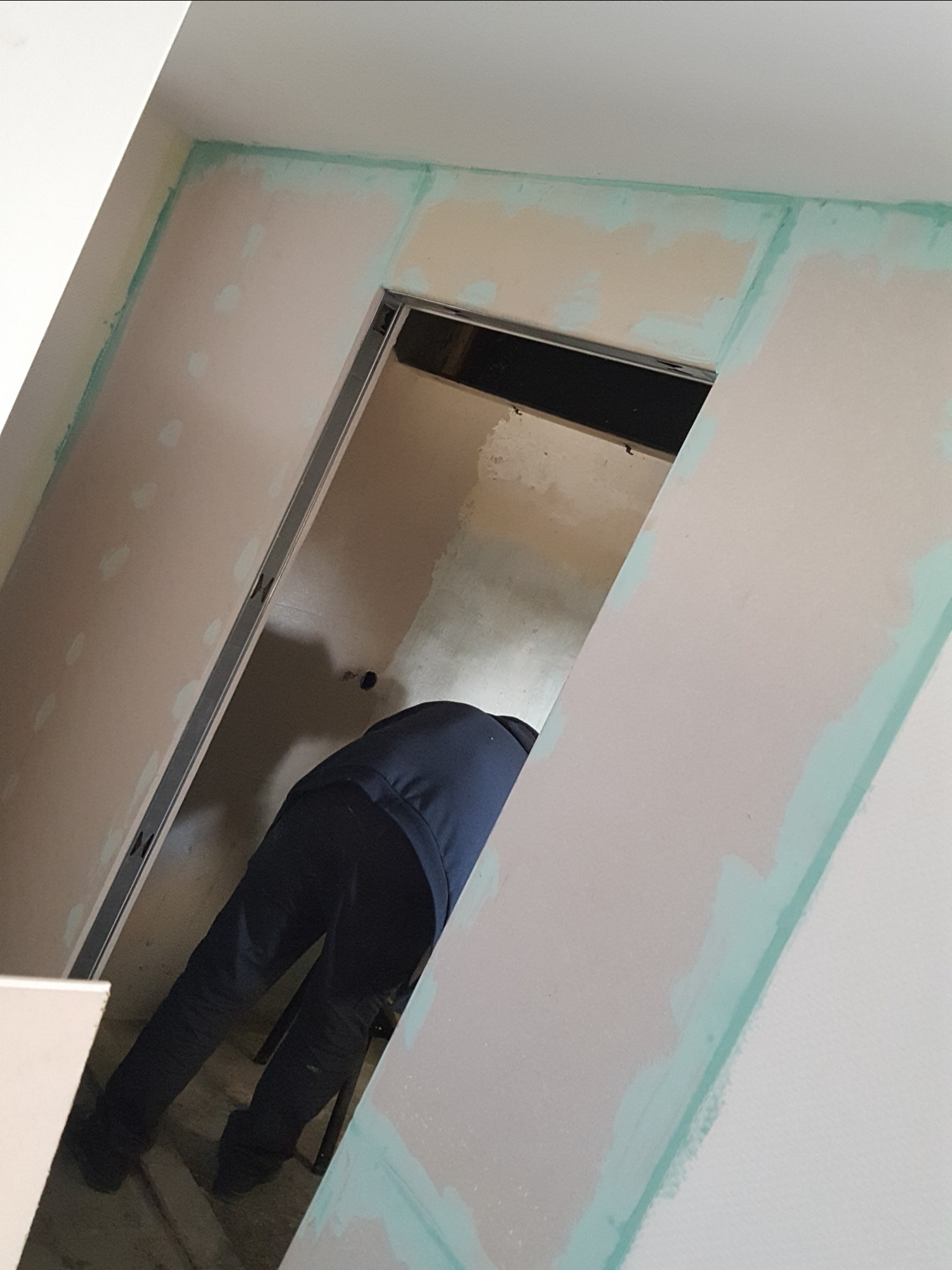 Pracownik w trakcie szpachlowania ścian w pomieszczeniu z metalową ościeżnicą, widoczne ślady szpachli i zielona taśma malarska na krawędziach.