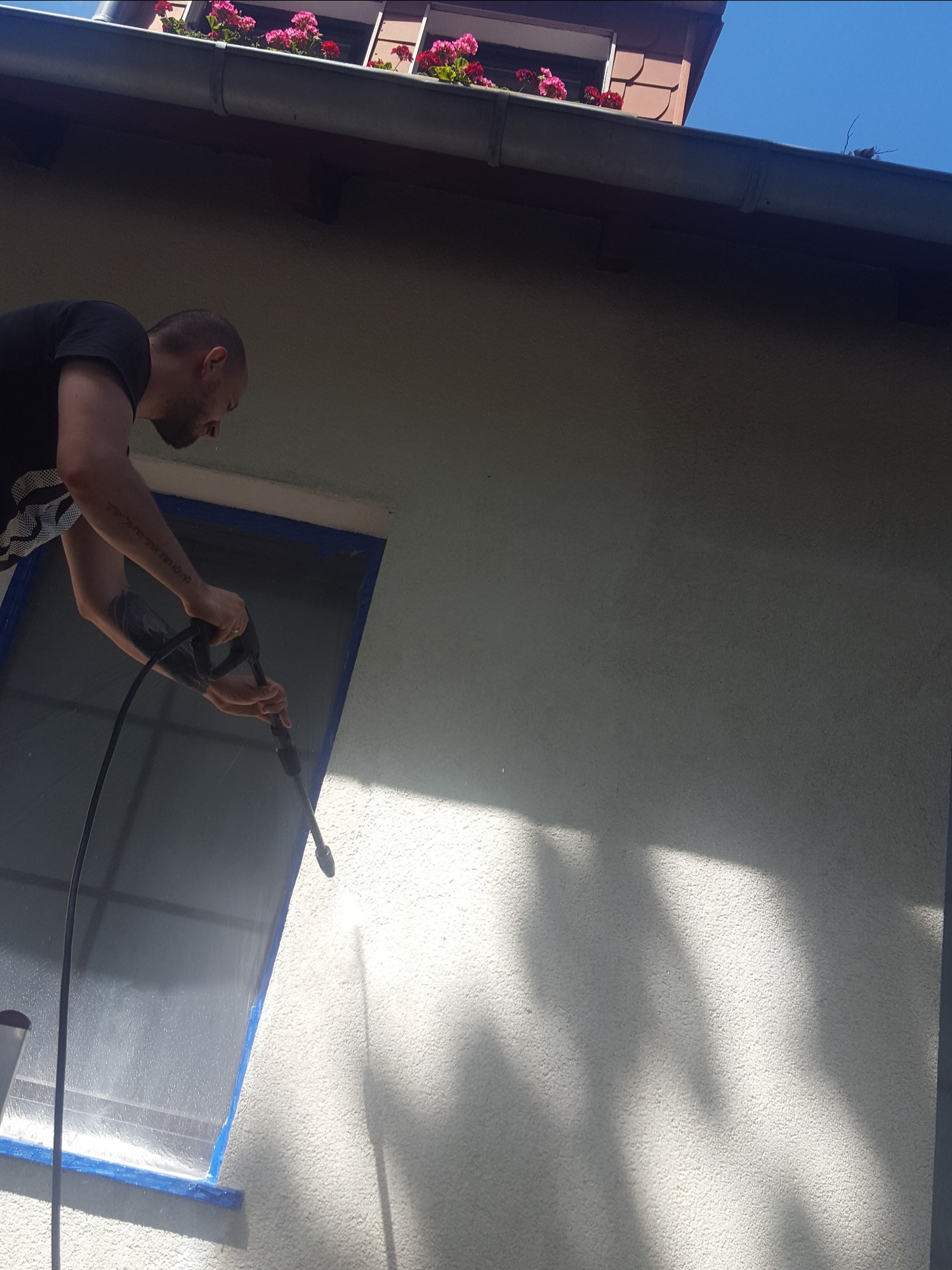 Mężczyzna z tatuażem na ramieniu czyści elewację domu myjką ciśnieniową, okno zabezpieczone niebieską taśmą malarską, w tle rynna z czerwonymi kwiatami.