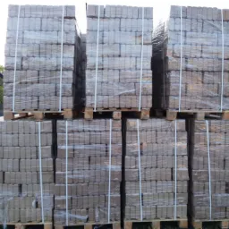 Ułożone na paletach i owinięte folią pakiety brykietu drzewnego, widoczne inne palety w tle.