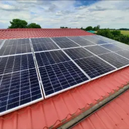 Panel fotowoltaiczny na czerwonym dachu z blachy trapezowej, widok z góry, w tle zielone drzewa i pochmurne niebo.