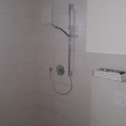 Haus Technik Instalacje Sanitarno Grzewcze - Nowoczesna kabina prysznicowa z chromowaną armaturą, białymi płytkami i widocznymi przewodami elektrycznymi na suficie, pudełko z akcesoriami marki Kludi na ścianie.