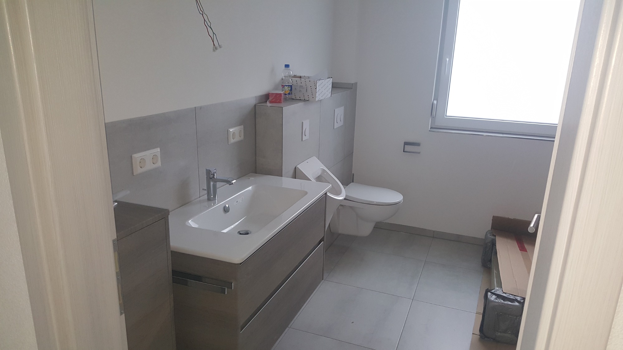 Nowoczesna łazienka w trakcie wykańczania: widoczna umywalka z szafką w kolorze drewna, podwieszana toaleta, szare płytki ścienne, wystające kable ze ściany i materiały budowlane na podłodze.