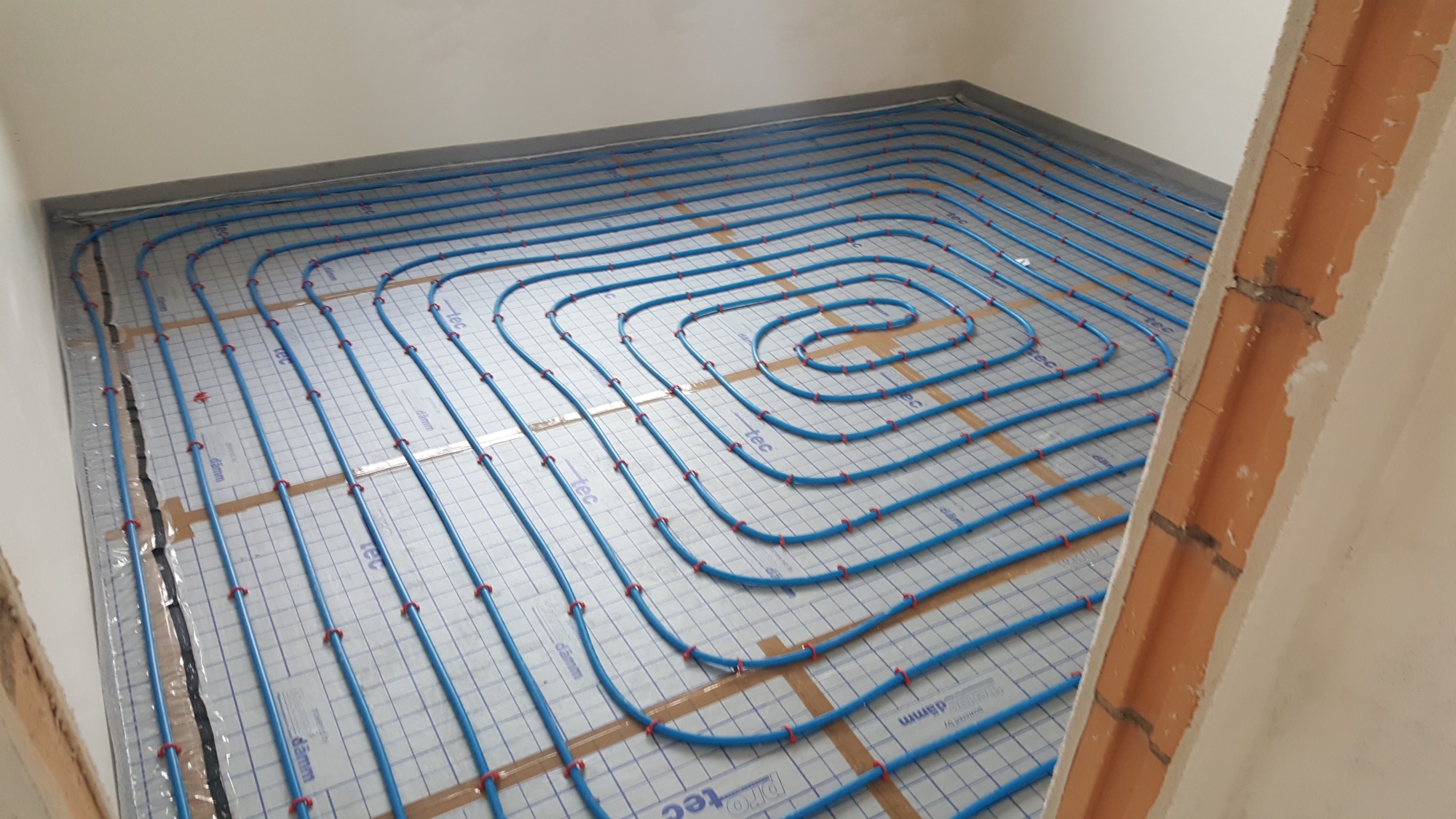 Niebieskie rury ogrzewania podłogowego ułożone spiralnie na izolacji z nadrukowaną siatką, mocowane czerwonymi klipsami, widok z góry w surowym pomieszczeniu.