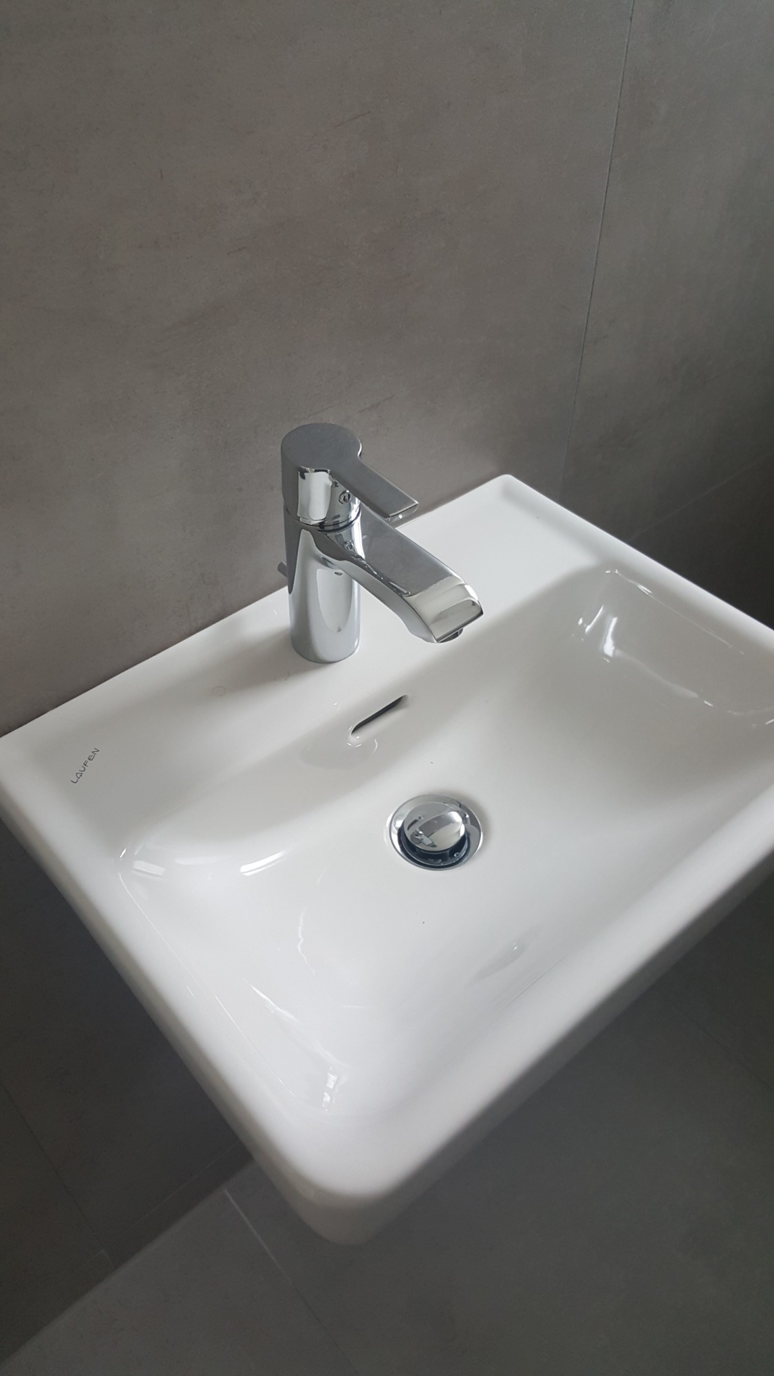 Biała, ceramiczna umywalka marki Laufen z chromowaną baterią i odpływem, zamontowana na tle szarej, matowej ściany.