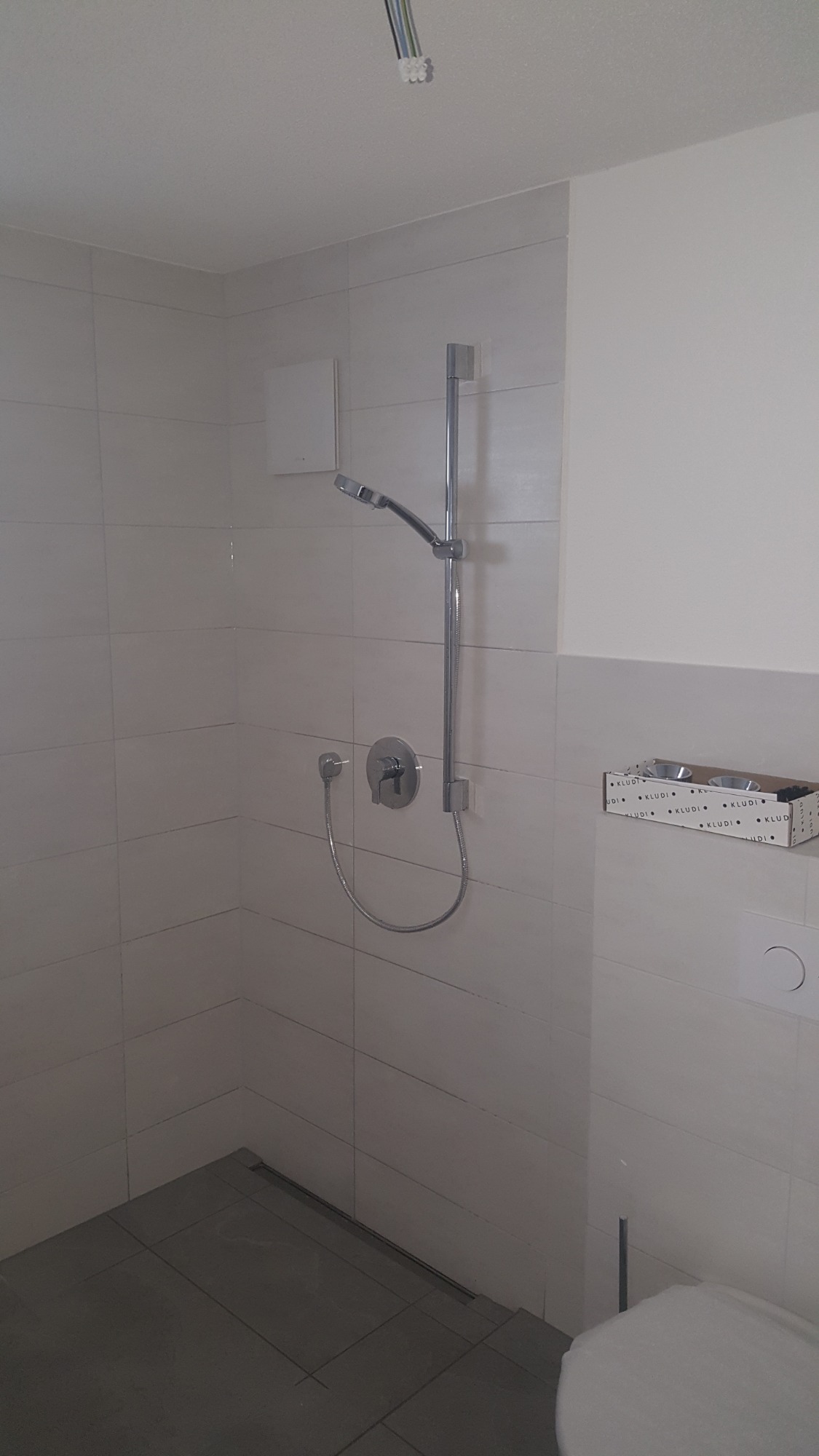 Nowoczesna kabina prysznicowa z chromowaną armaturą, białymi płytkami i widocznymi przewodami elektrycznymi na suficie, pudełko z akcesoriami marki Kludi na ścianie.