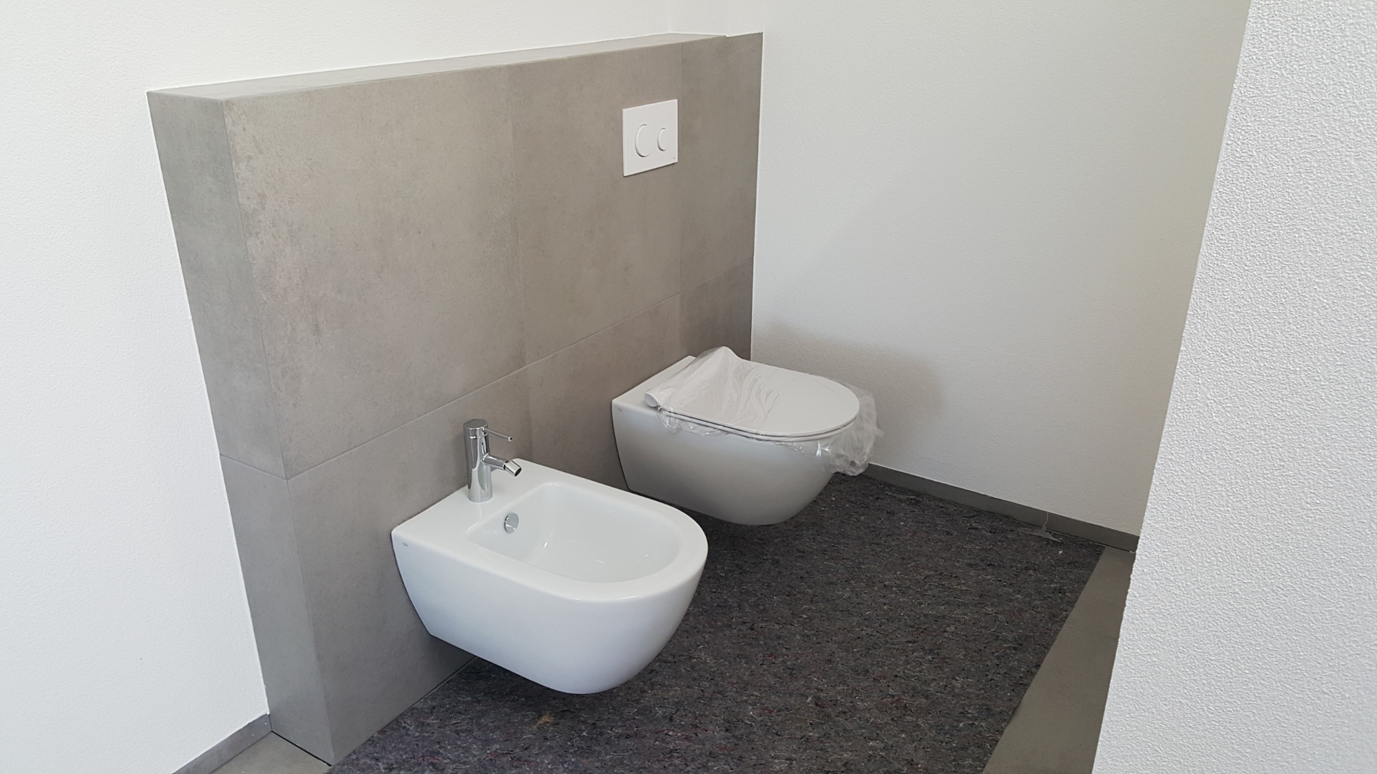 Nowoczesna łazienka z białym bidetem i podwieszaną toaletą, oba urządzenia ceramiczne, zamontowane na tle ściany wyłożonej szarymi płytkami, toaleta zabezpieczona folią ochronną, widoczne przyciski...