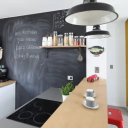 Nowoczesna kuchnia z czarną ścianą tablicową z napisem, drewnianym blatem, białymi szafkami, wiszącymi lampami w stylu industrialnym, i czerwonym stołkiem barowym.