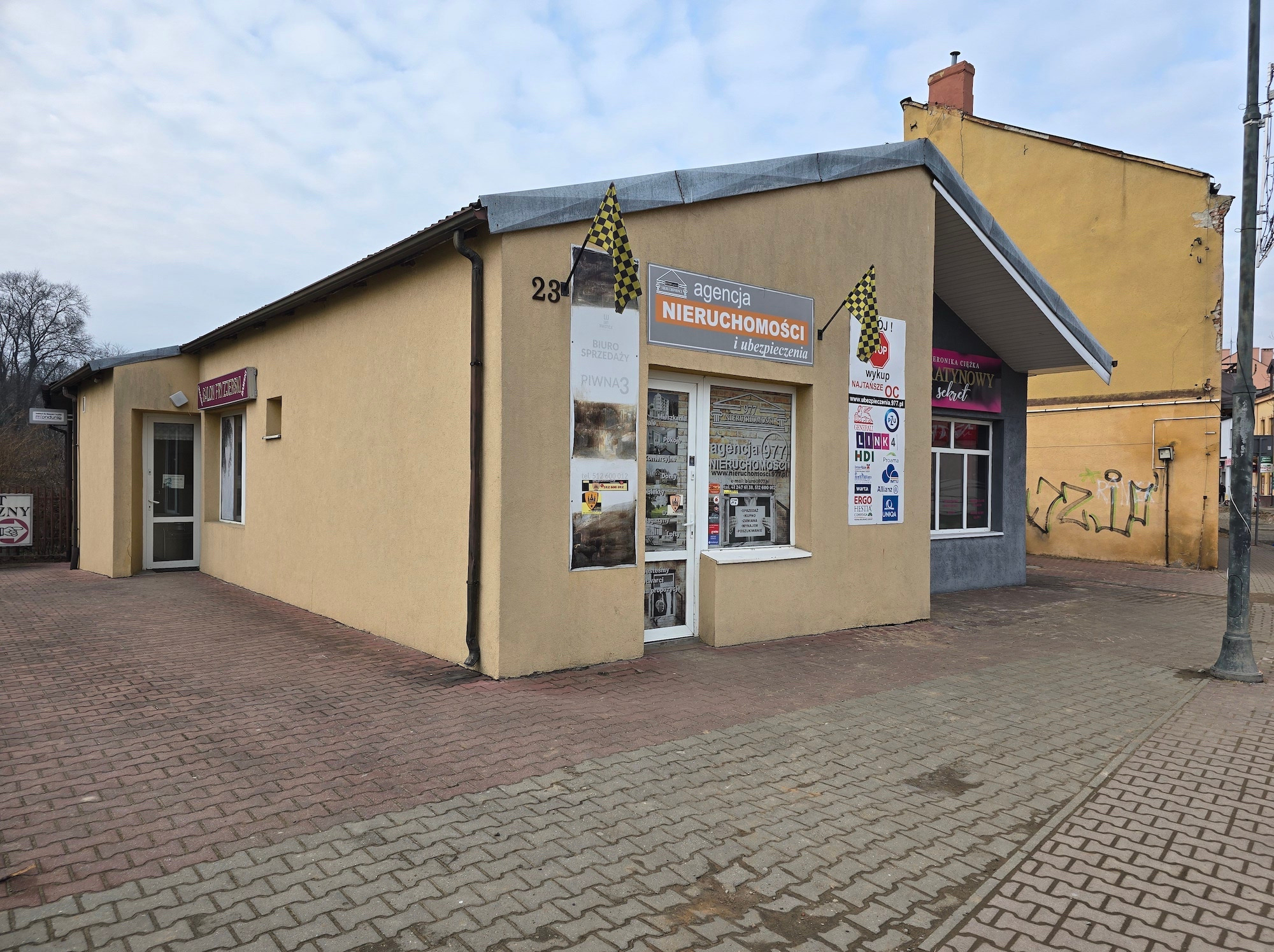 Fasada biura agencji nieruchomości z szyldem 'Agencja Nieruchomości i ubezpieczenia' oraz banerami reklamowymi. Widok na budynek z chodnika.