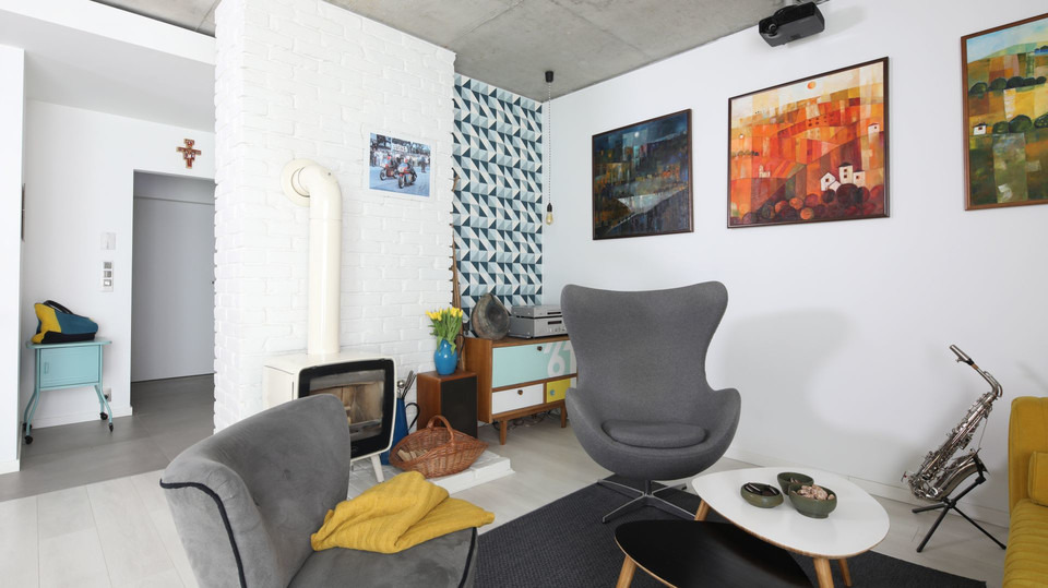 Jasne wnętrze salonu z białymi ścianami, kominkiem kaflowym, szarymi fotelami typu 'Egg' i saksofonem na statywie, oświetlone światłem dziennym i sztucznym, z dekoracyjnymi obrazami na ścianie.