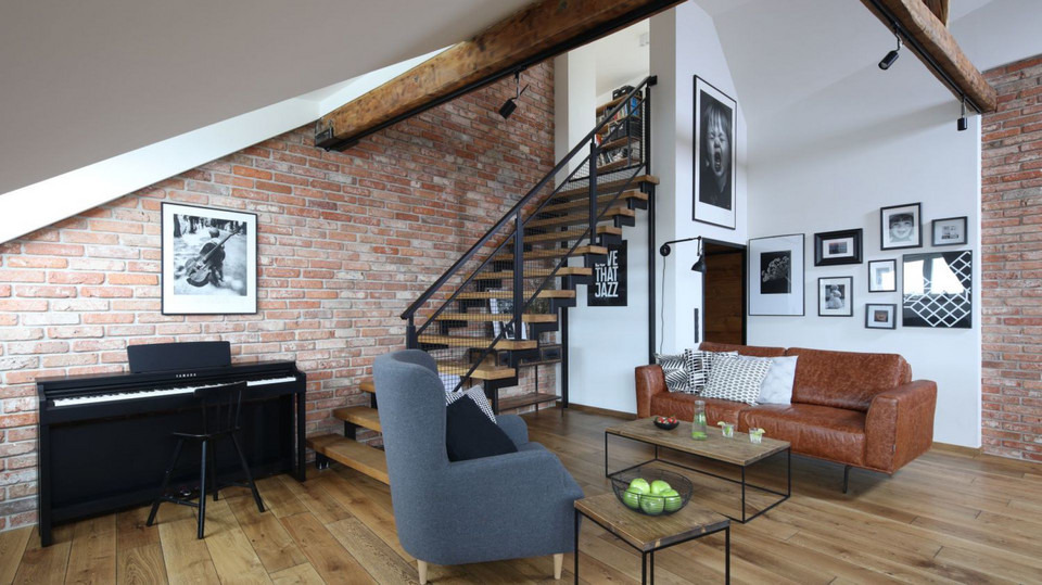 Loftowe wnętrze z ceglaną ścianą, drewnianymi belkami, schodami z metalową balustradą, pianinem Yamaha i galerią czarno-białych zdjęć nad brązową skórzaną sofą.