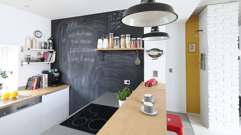 Nowoczesna kuchnia z czarną ścianą tablicową z napisem, drewnianym blatem, białymi szafkami, wiszącymi lampami w stylu industrialnym, i czerwonym stołkiem barowym.