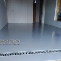 Świeżo wylana, szara podłoga z odpływem liniowym w surowym wnętrzu z widocznym nagrzewnicą w ścianie.