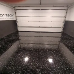Błyszcząca, czarna posadzka z białymi drobinkami w garażu z otwartą, segmentową bramą garażową i logo firmy Epoxytech na ścianie.