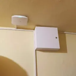 Centrala alarmowa w białej obudowie i czujnik na suficie, widoczne na tle żółtej ściany i sufitu, obok piłki do ćwiczeń.